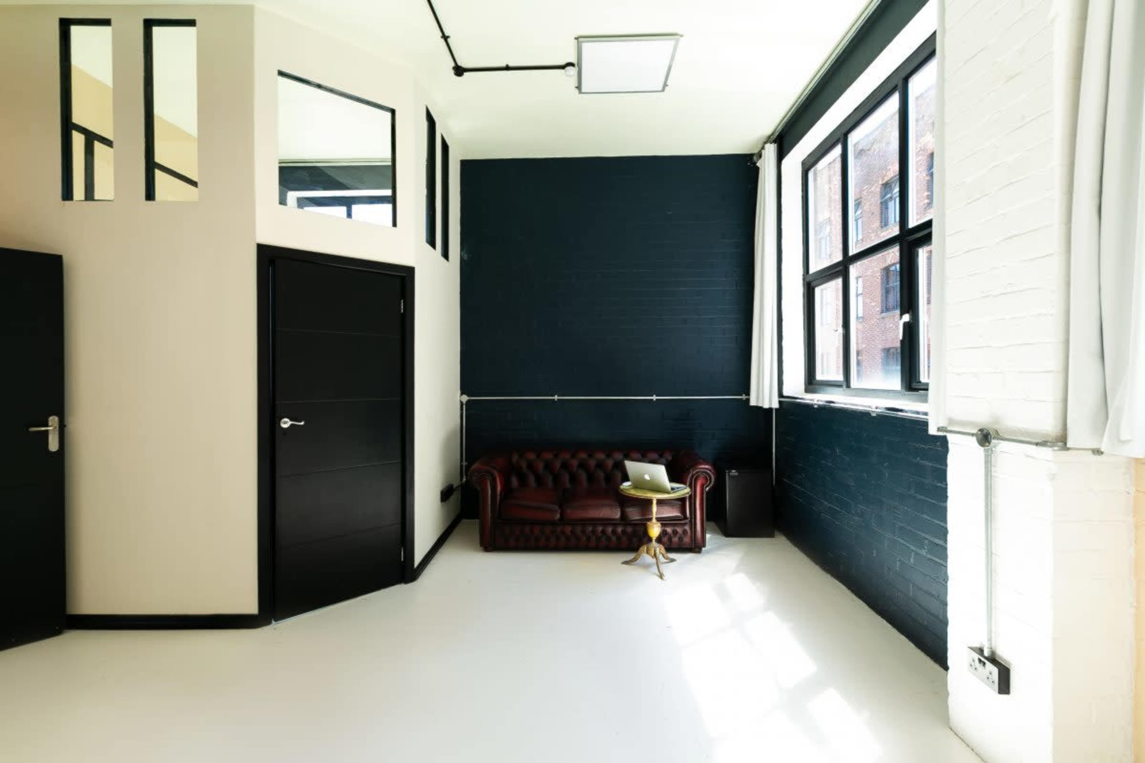 A spacious interior room with a dark wall, a brown chesterfield sofa, a small side table, and large windows allowing natural light to enter.