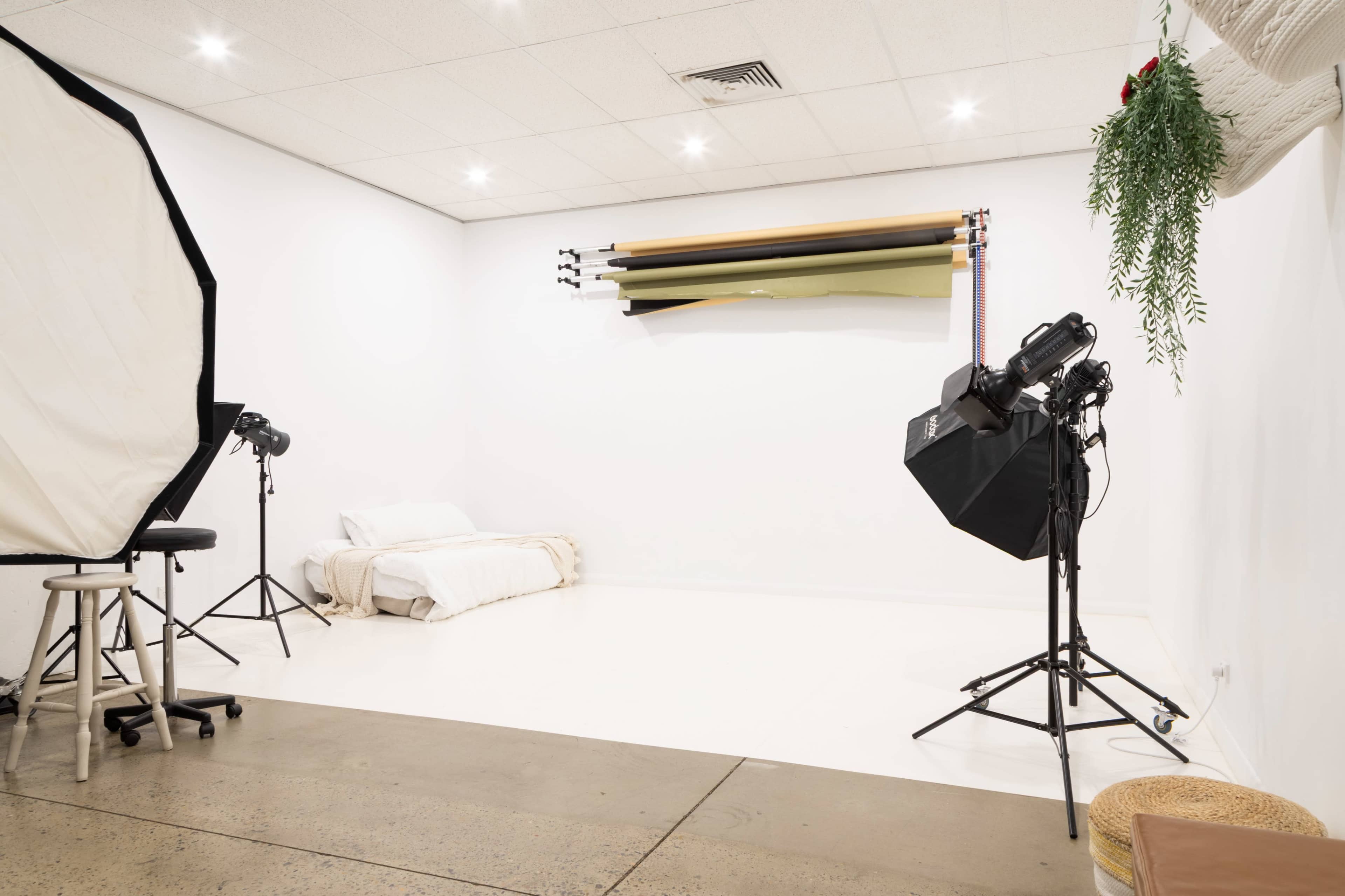 The image shows a photography studio featuring a simple bed against a white wall, with backdrops and studio lighting equipment arranged on either side.
