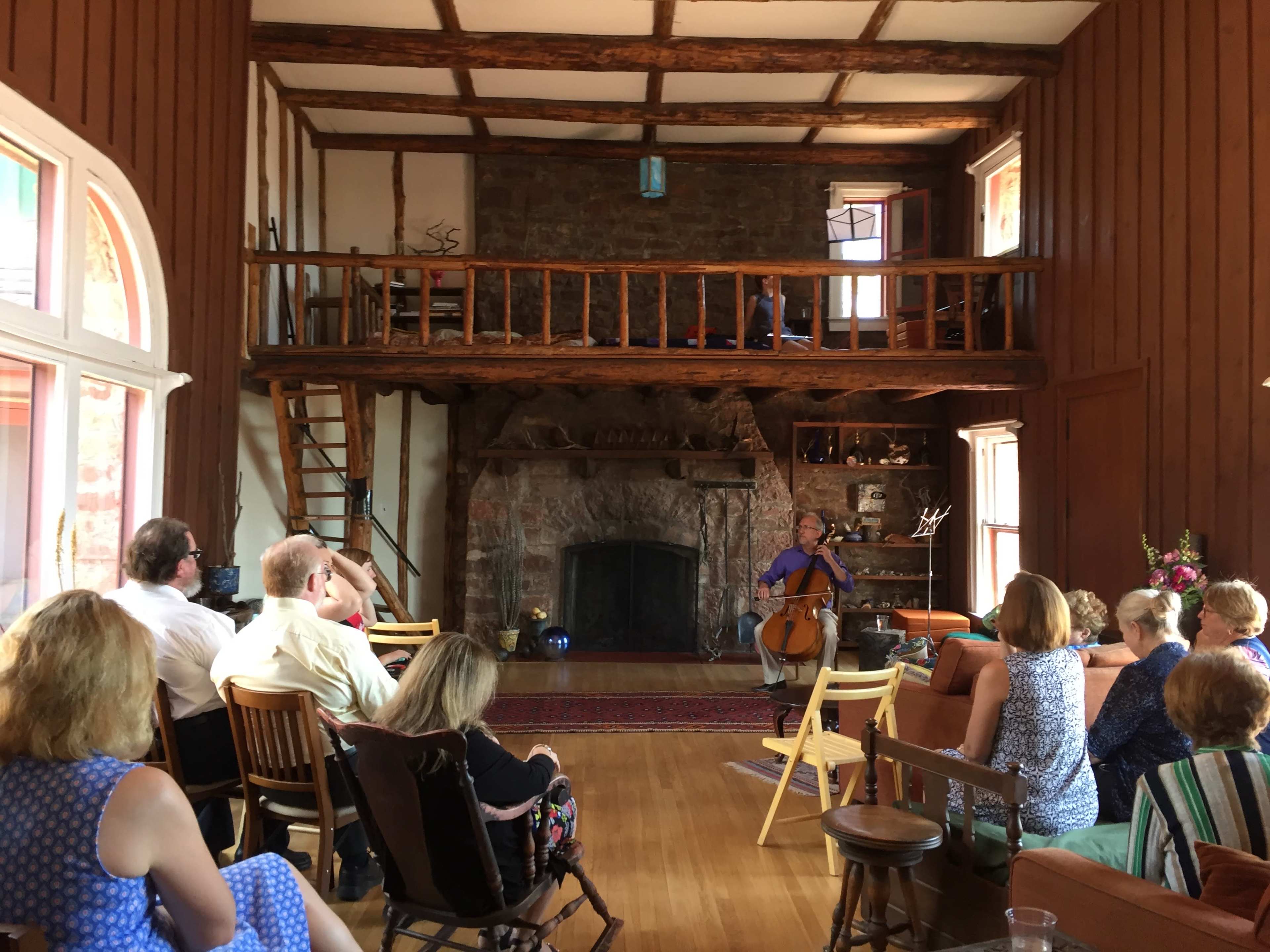 A cellist performs in a spacious, wooden-beamed room with an audience seated on various chairs.