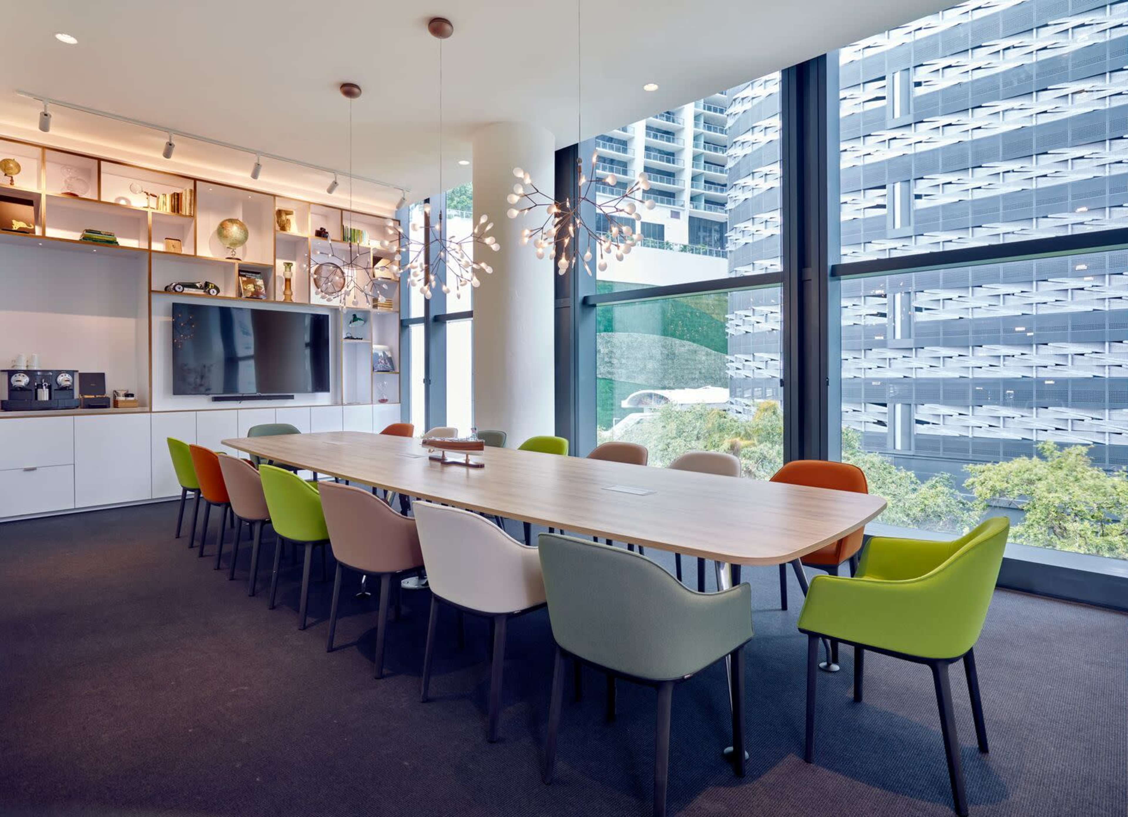 A long table with variously colored chairs is set up in a modern conference room with large windows overlooking a courtyard and building structure.