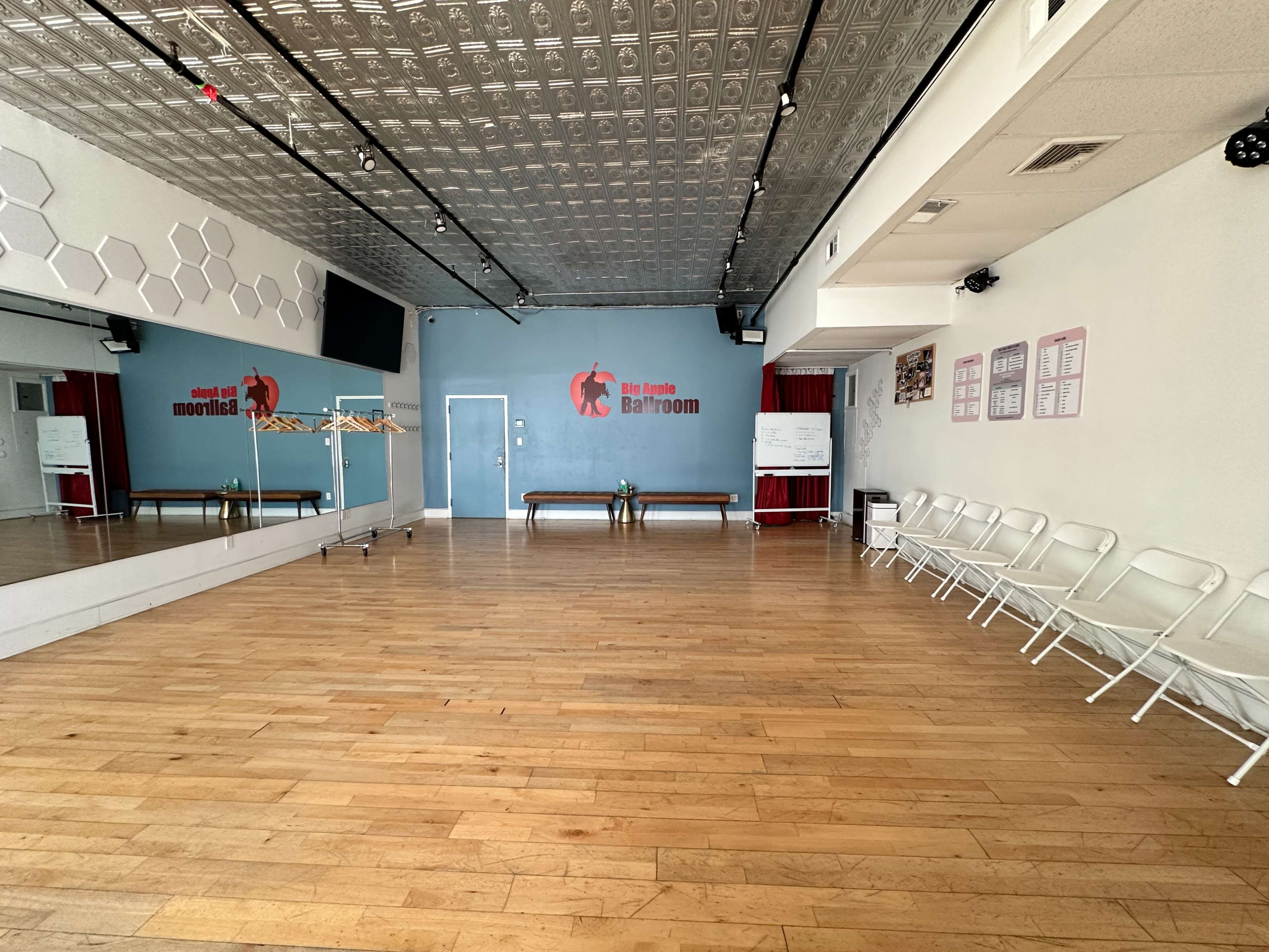 The image shows a spacious dance studio with hardwood flooring, a mirrored wall, and red accents, including a logo on the wall.
