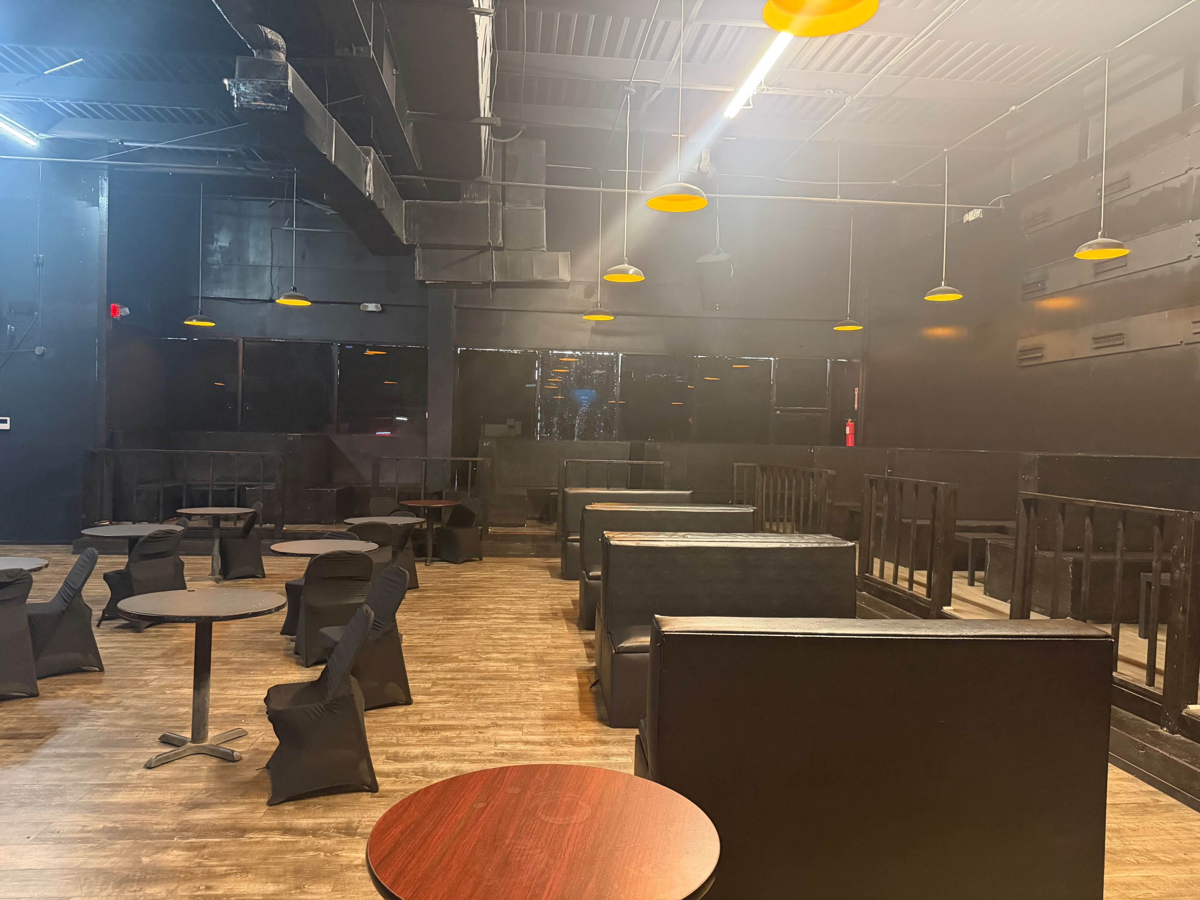 The image shows a dimly lit restaurant or lounge interior featuring several black tables and booths arranged throughout the space.