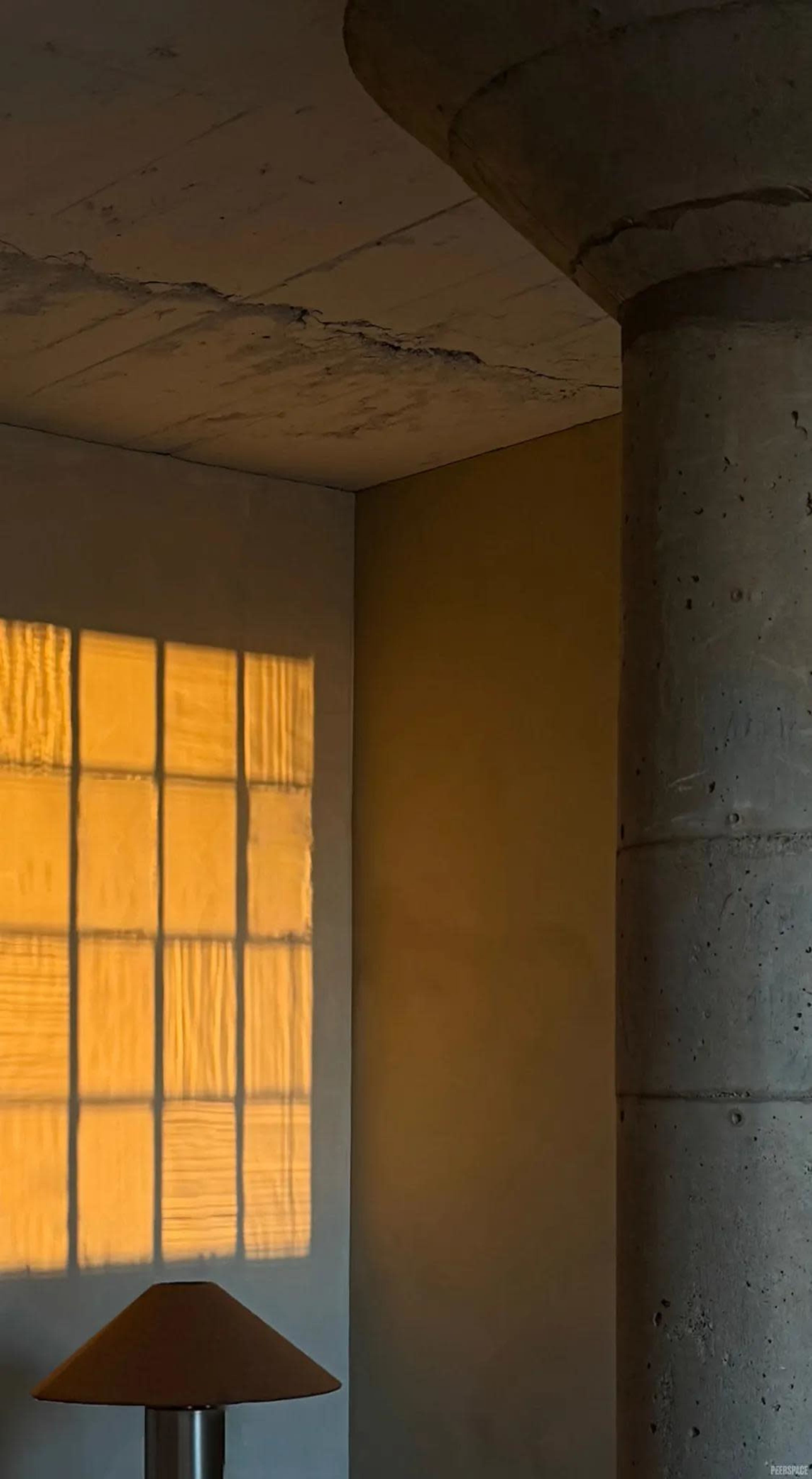 A warm yellow light casts a rectangular pattern on a wall from a window, highlighting a concrete column and a lamp in the corner of a room.