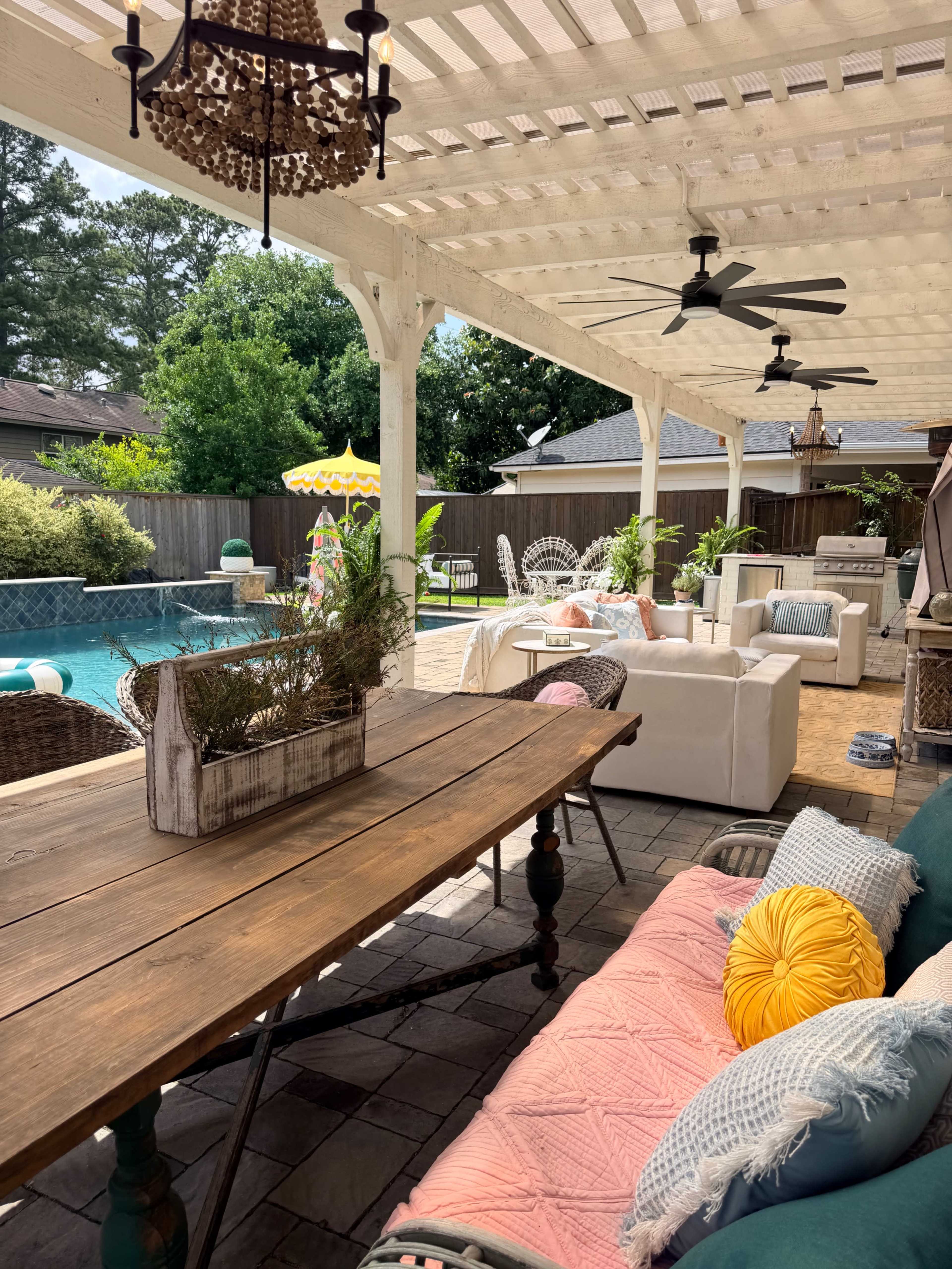 The image shows a patio area with a wooden table, comfortable seating, and a swimming pool surrounded by greenery.