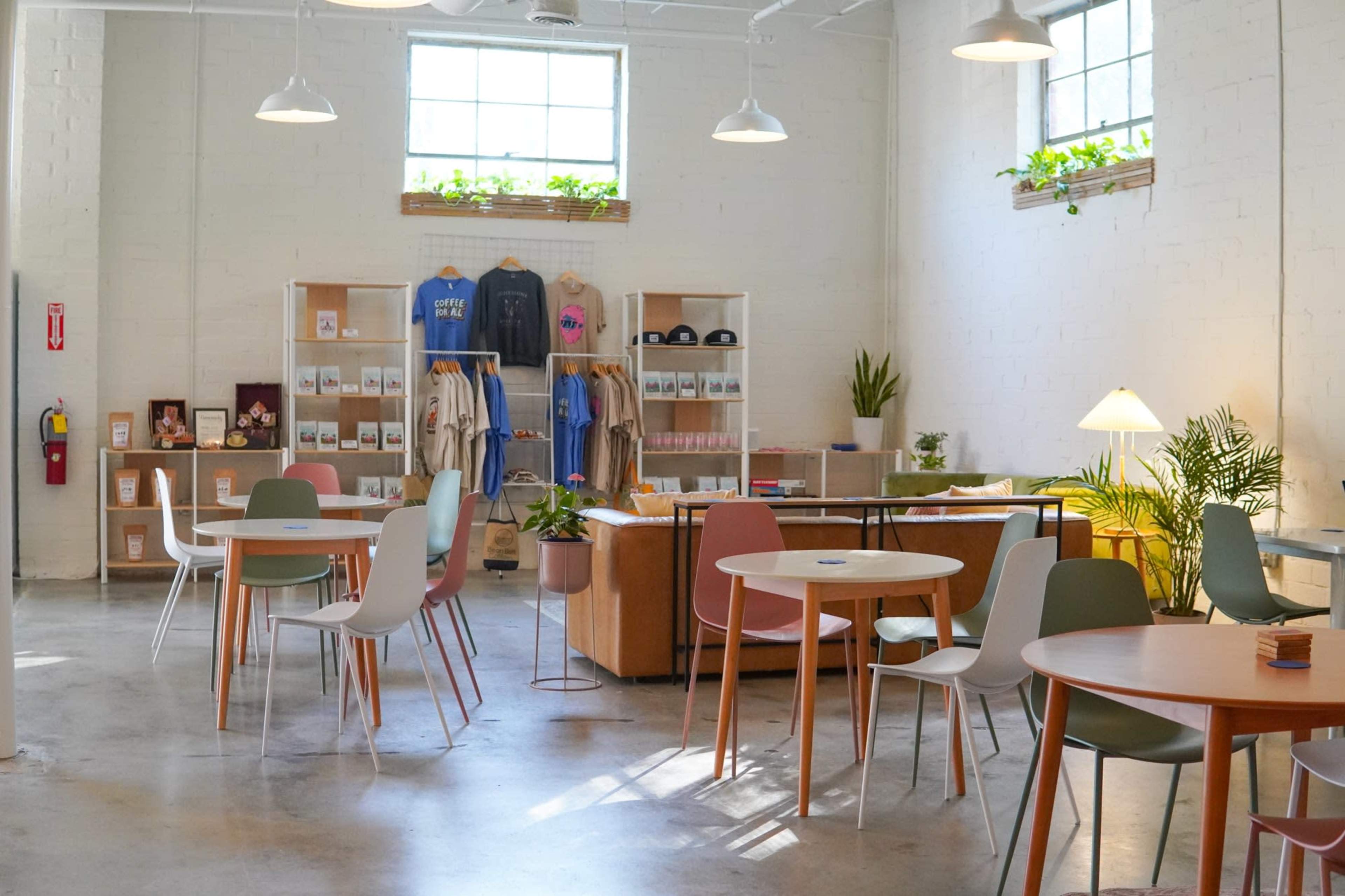 A bright, modern cafe features various seating arrangements, merchandise displays, and plants against a light-colored brick background.
