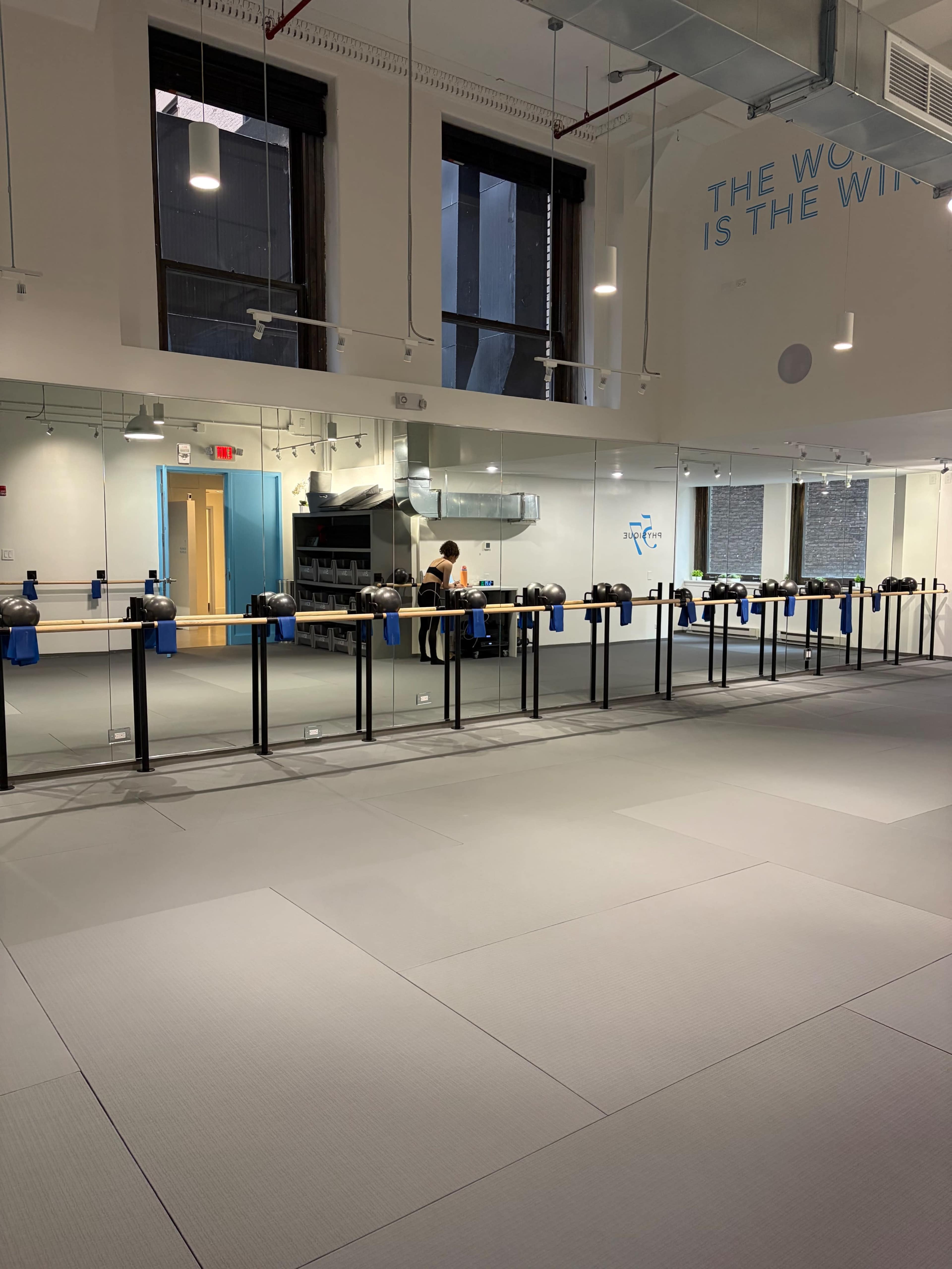 The image shows a spacious, well-lit studio with gray flooring, multiple black barre stations, and a person preparing equipment at the back.