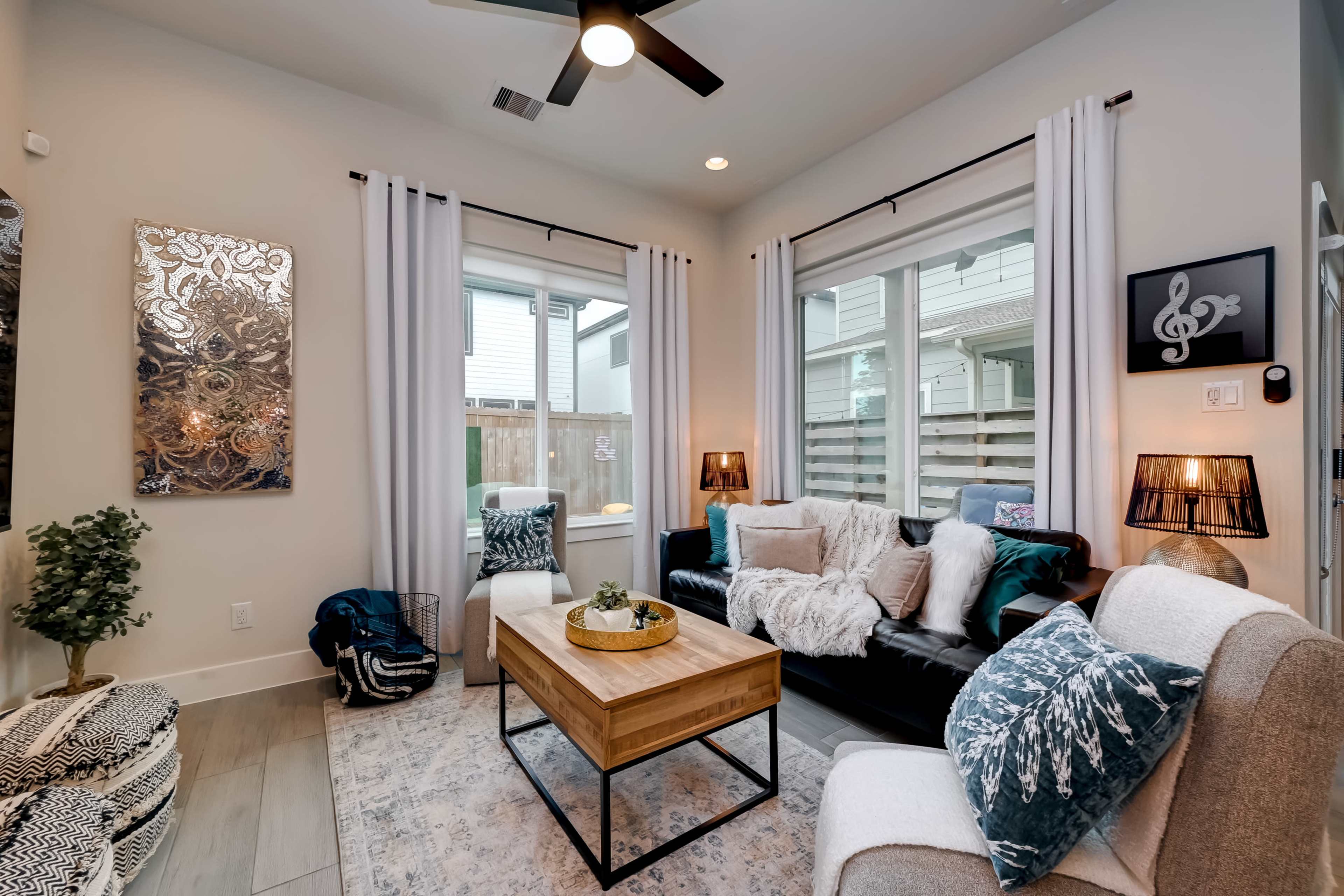 A modern living room features a black sofa, a wooden coffee table, and large windows with white curtains, decorated with plants and a textured area rug.