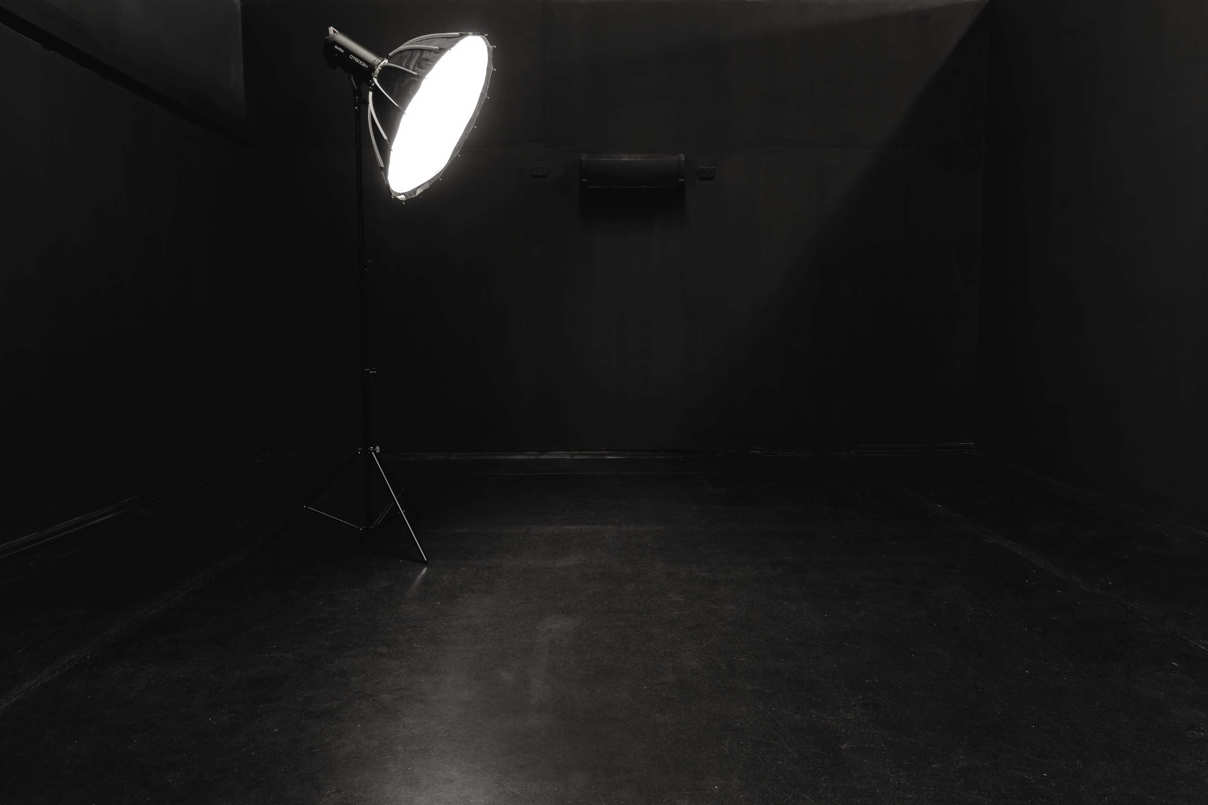 The image shows a dimly lit black room with a large spotlight on a stand illuminating part of the floor.