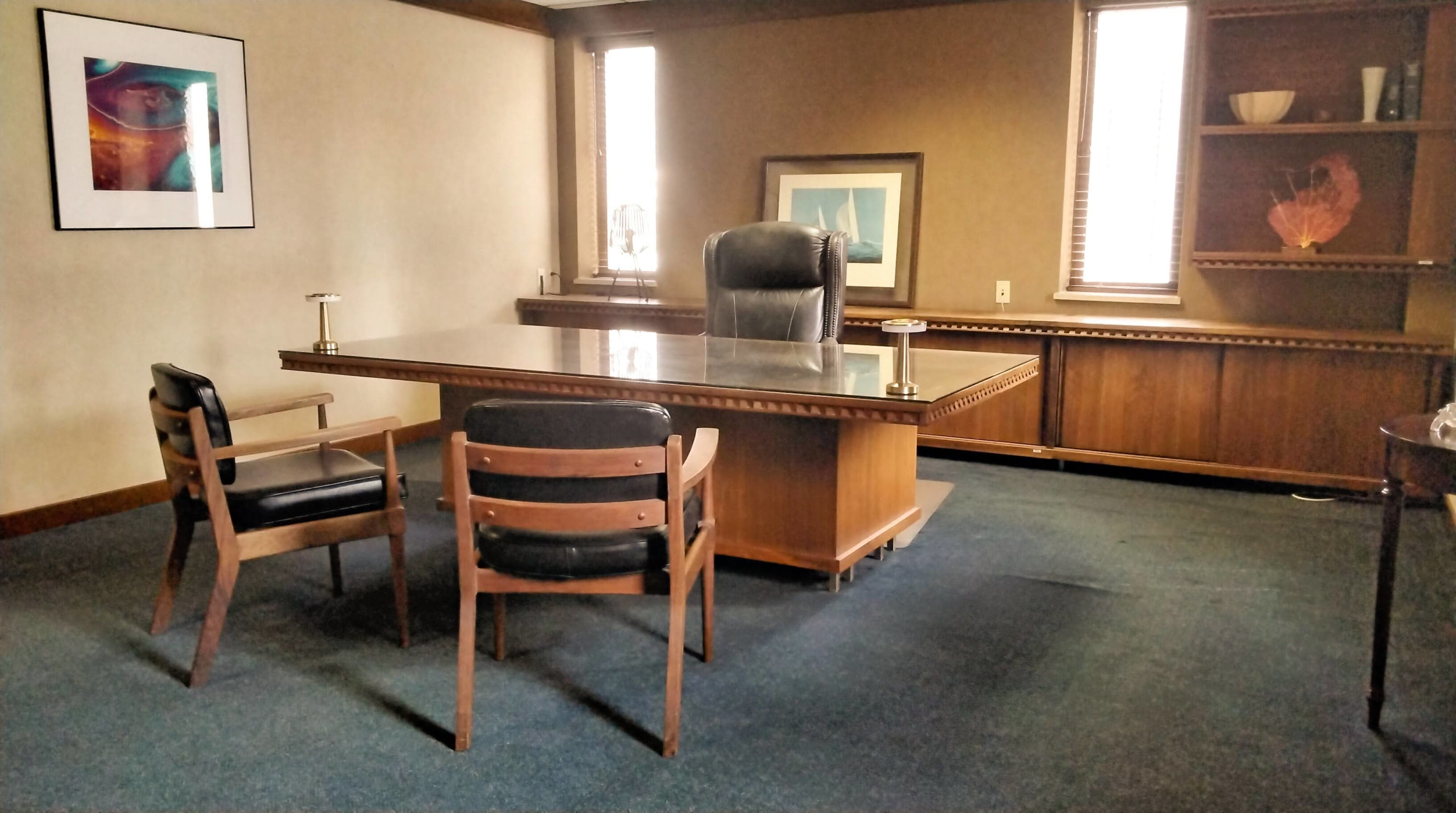 The image shows an office space featuring a large wooden desk, a leather office chair behind it, two wooden chairs in front, and minimal decor on the walls and shelves.