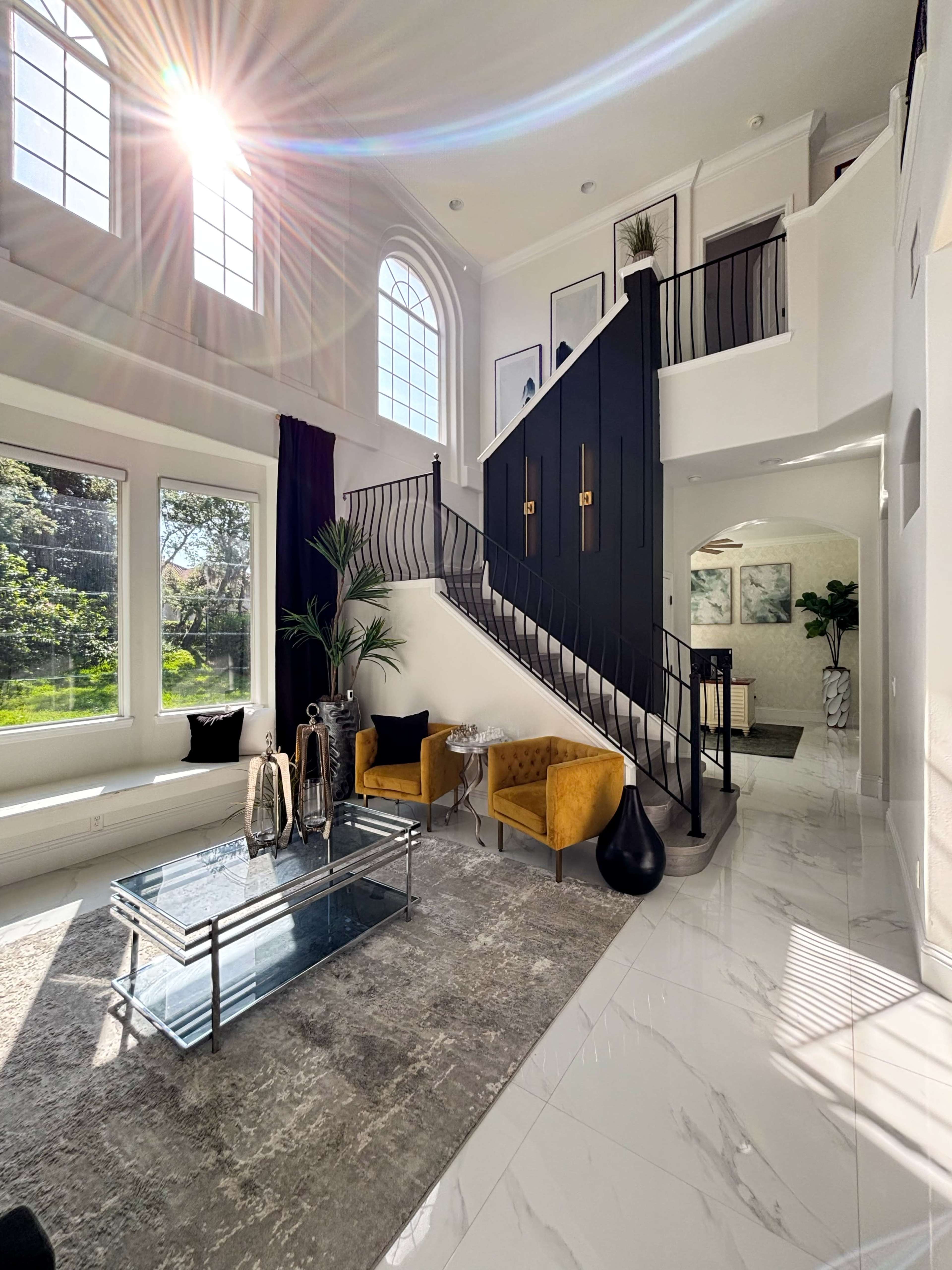 The image shows a bright foyer with a winding staircase, large windows, and modern furnishings, including yellow accent chairs and a glass coffee table.