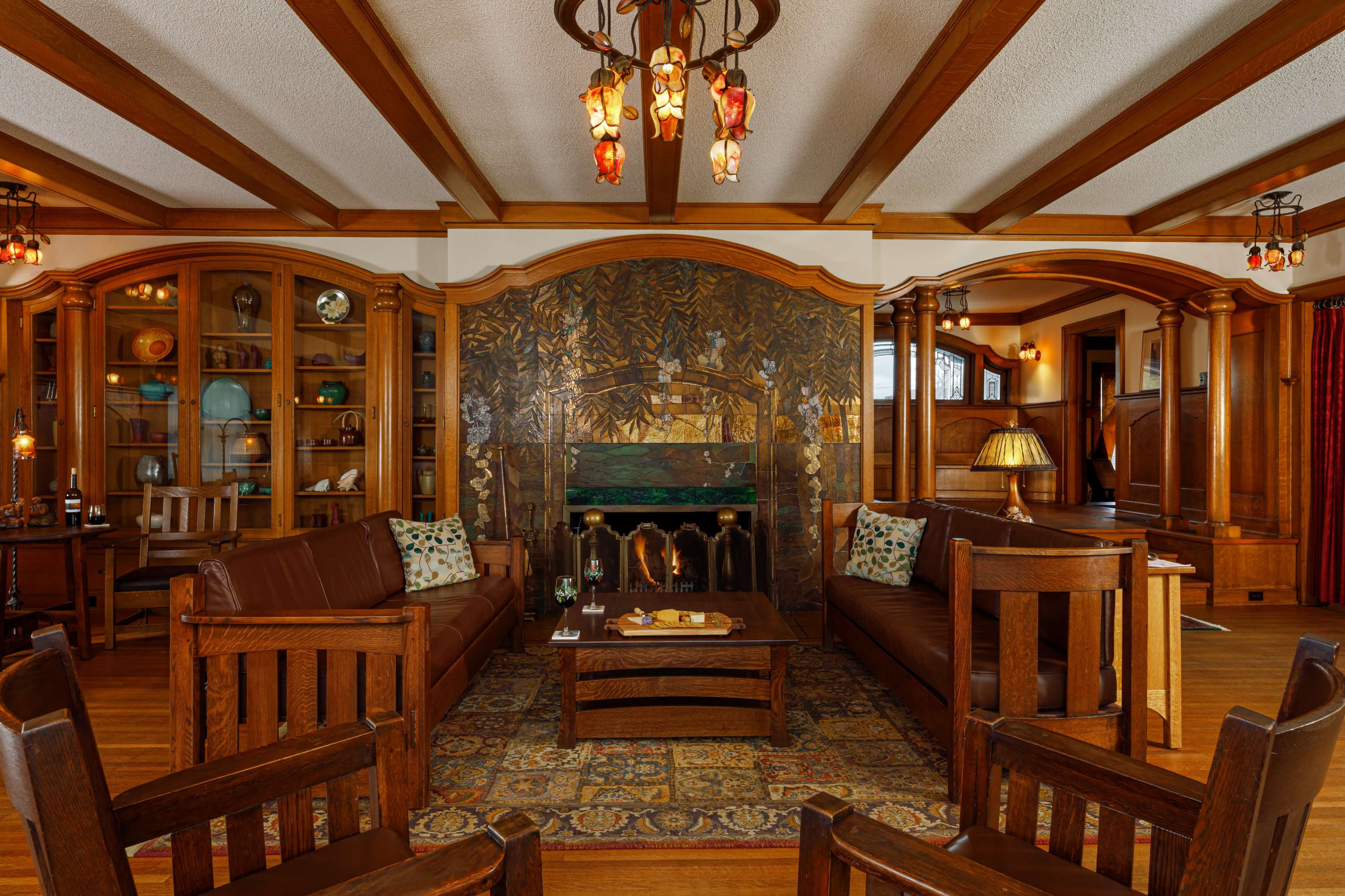 A cozy living room with wooden beams, two brown leather sofas facing a coffee table, and a large ornate fireplace decorated with a mural.