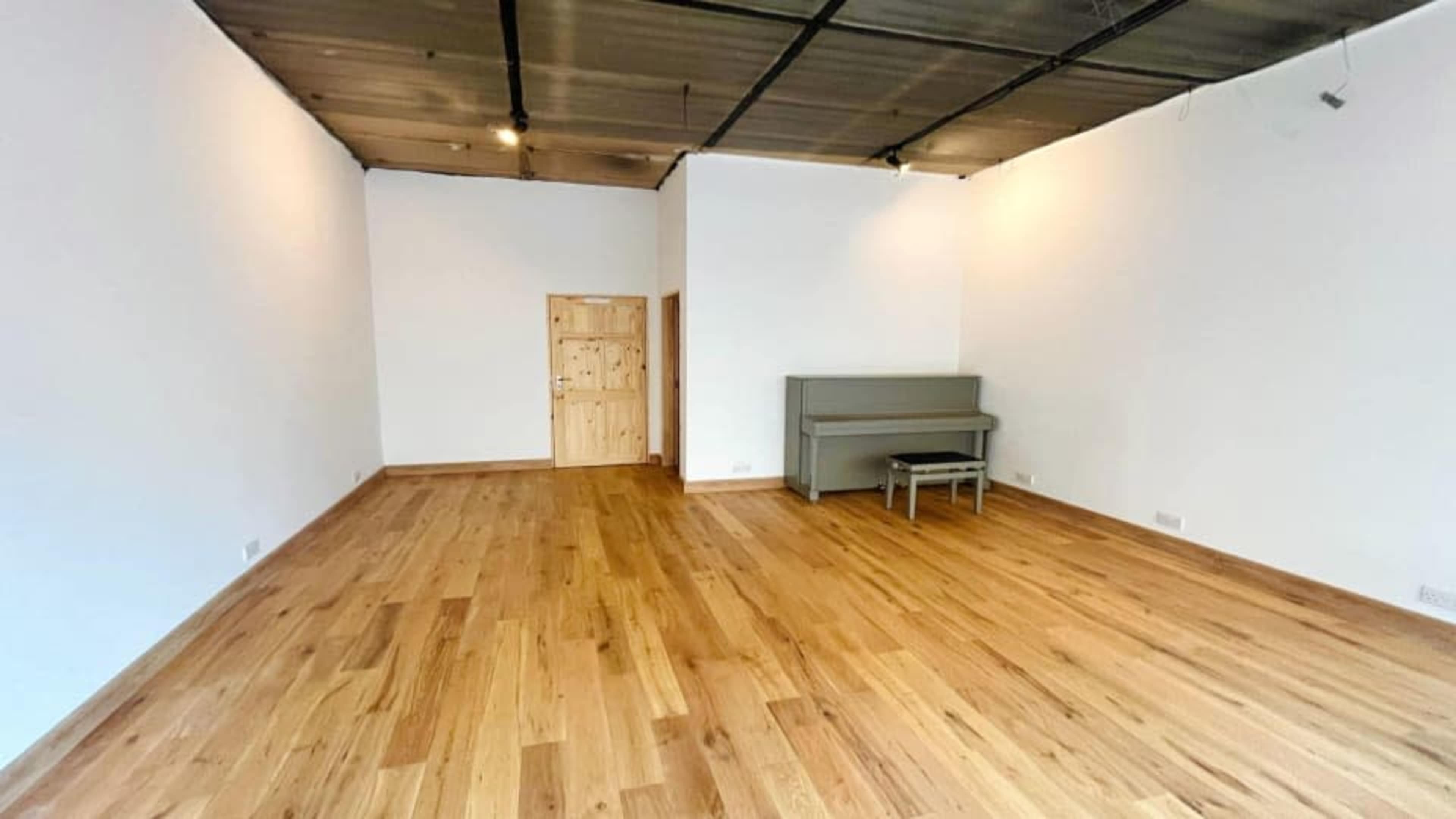 The image shows a spacious, empty room with wooden floors, a gray piano against one wall, and a light-colored door.