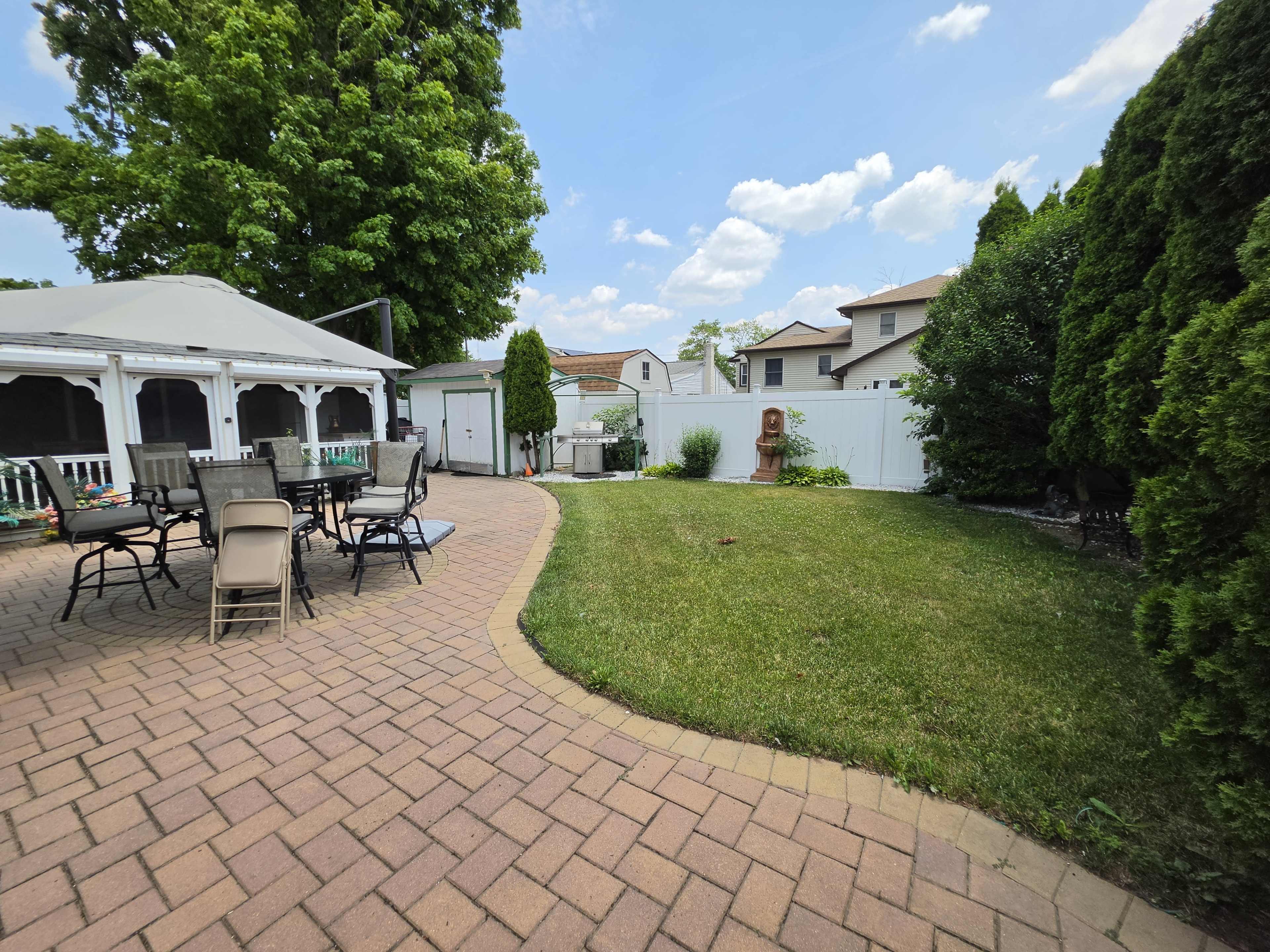 The image shows a landscaped backyard with a patio area featuring a table and chairs, surrounded by greenery and a fence.