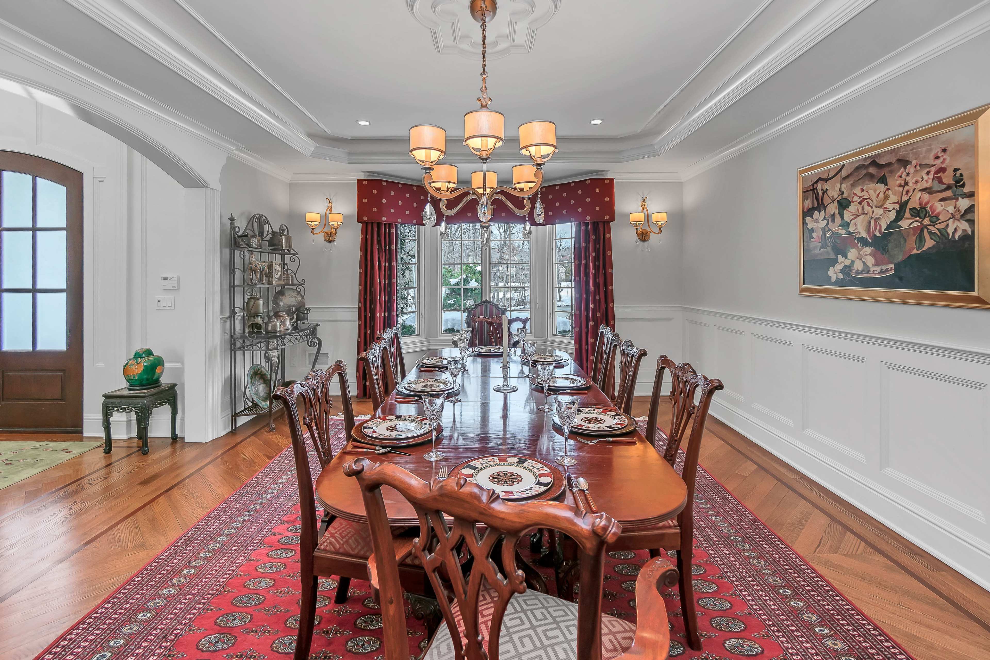 A formal dining room with a long wooden table set for a meal, surrounded by elegant chairs, and decorated with a patterned area rug and framed artwork on the walls.
