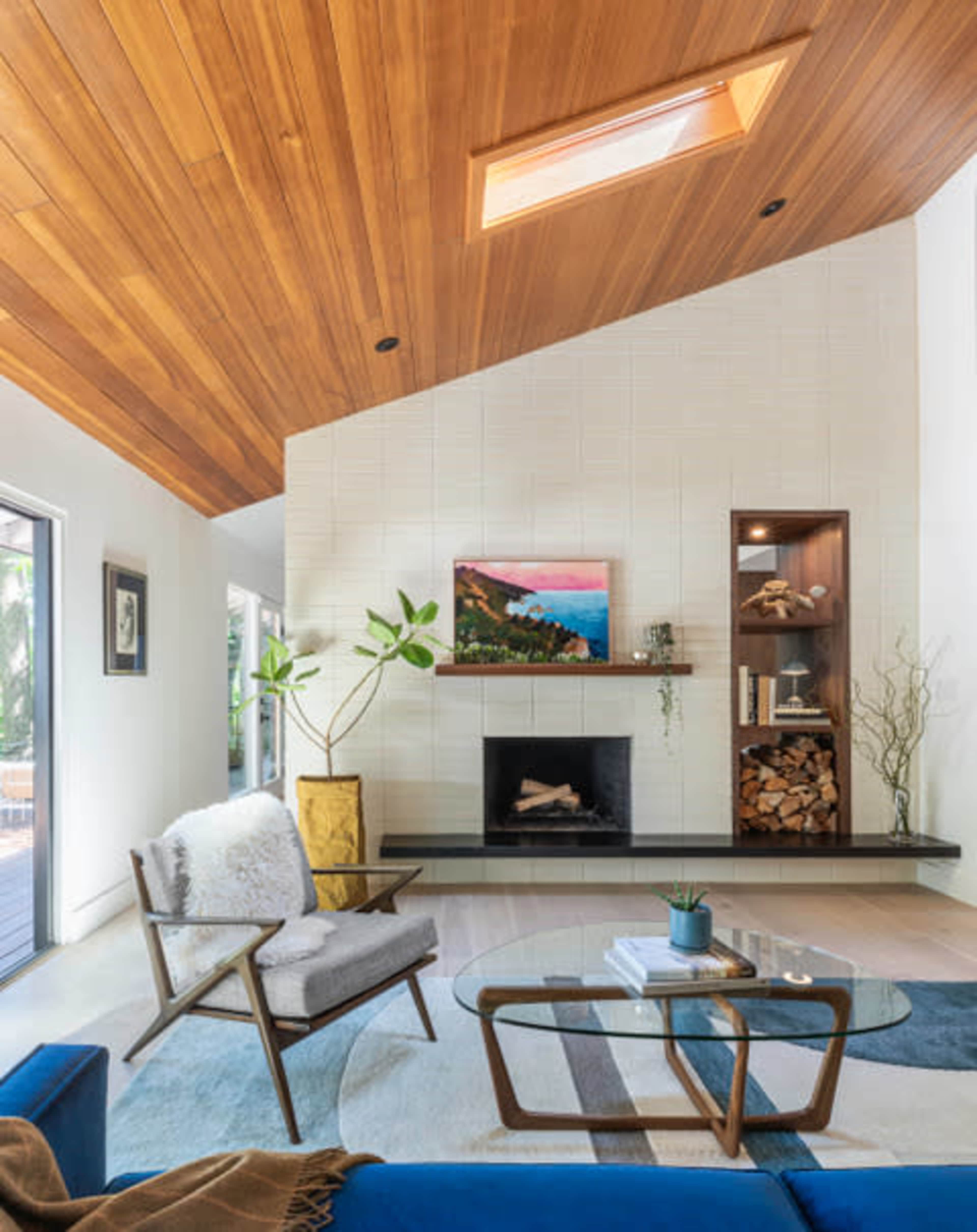A modern living room featuring a wooden ceiling, a fireplace with a mounted painting above it, and a combination of plants and decorative elements.