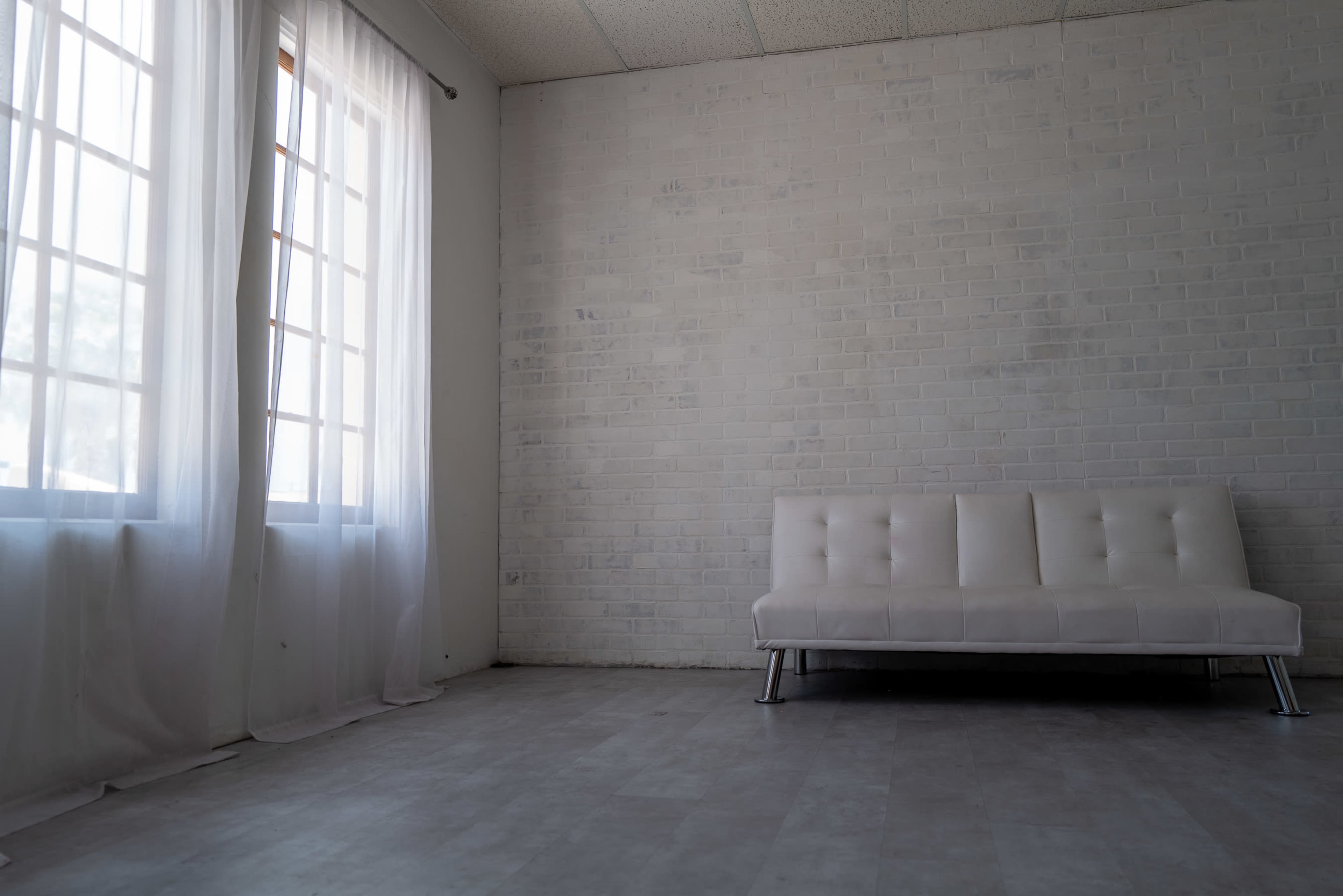 A minimalist room with a white brick wall, a white sofa, and large windows draped with sheer curtains.