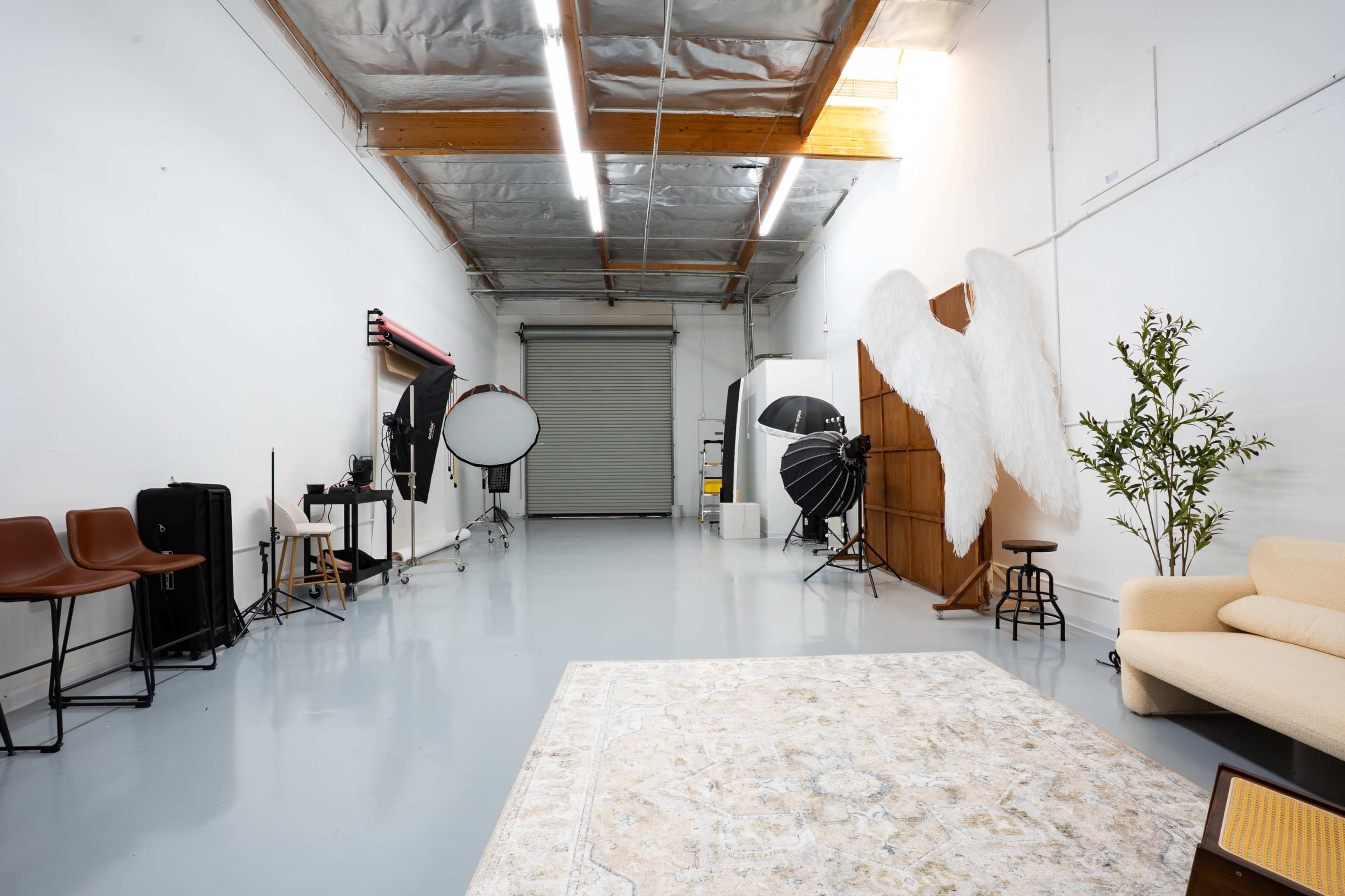 The image shows a spacious photography studio featuring a seating area, various lighting equipment, and a large pair of angel wings displayed on the wall.
