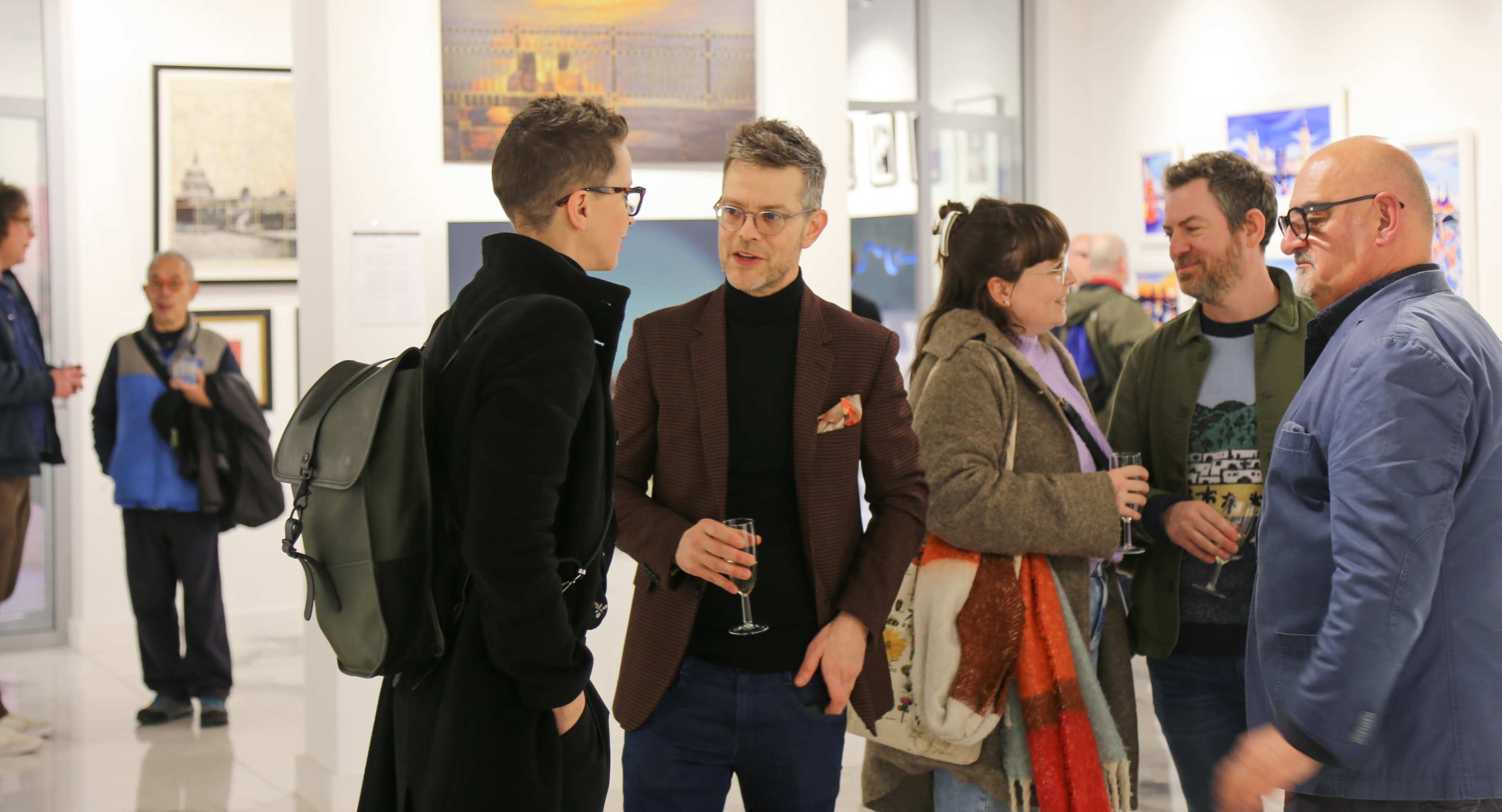 A group of people is engaged in conversation at an art gallery event, with various artworks displayed on the walls.