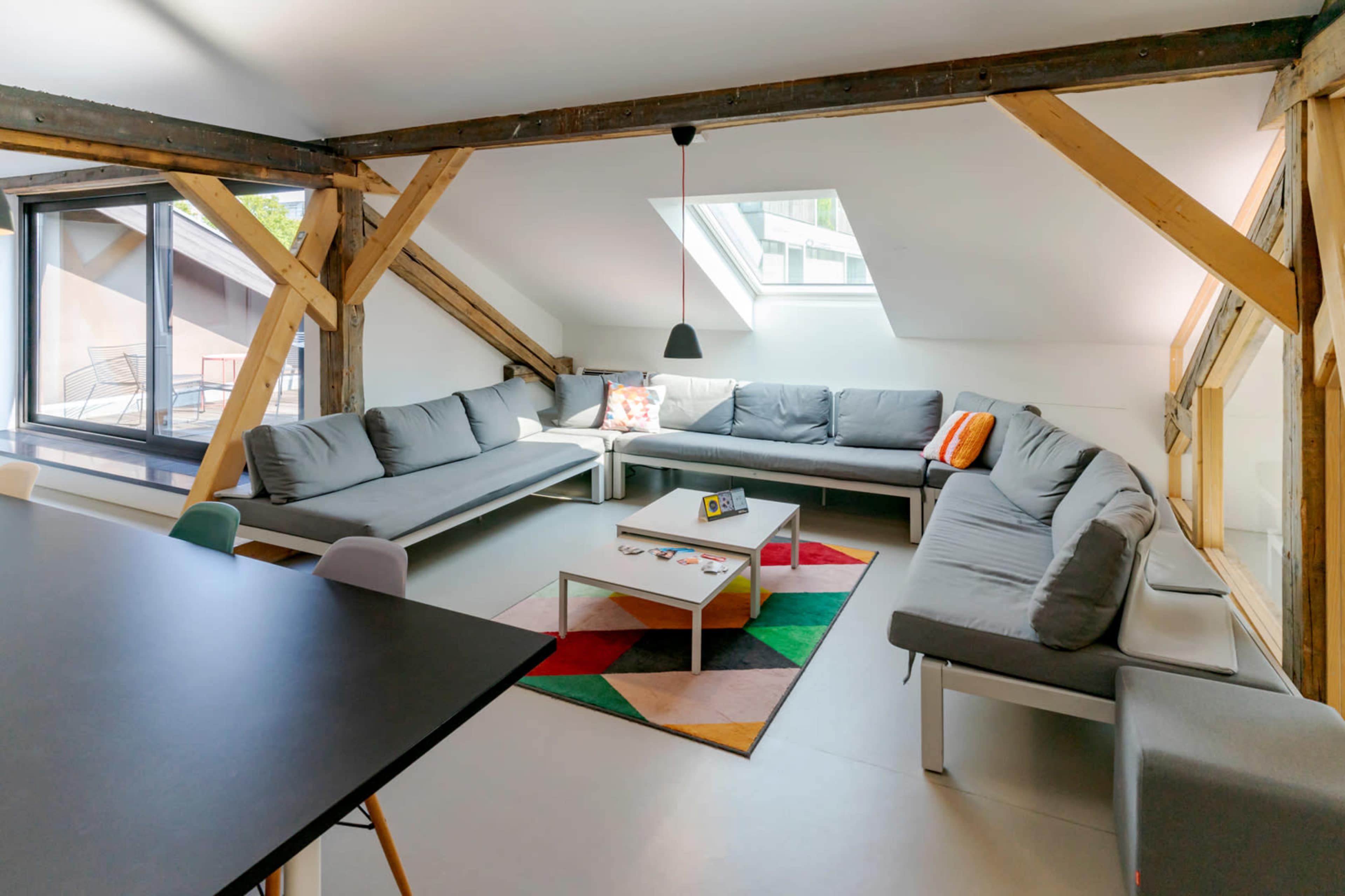 The image shows a modern, spacious living area with a large sectional sofa, a coffee table, and wooden beams in the ceiling.