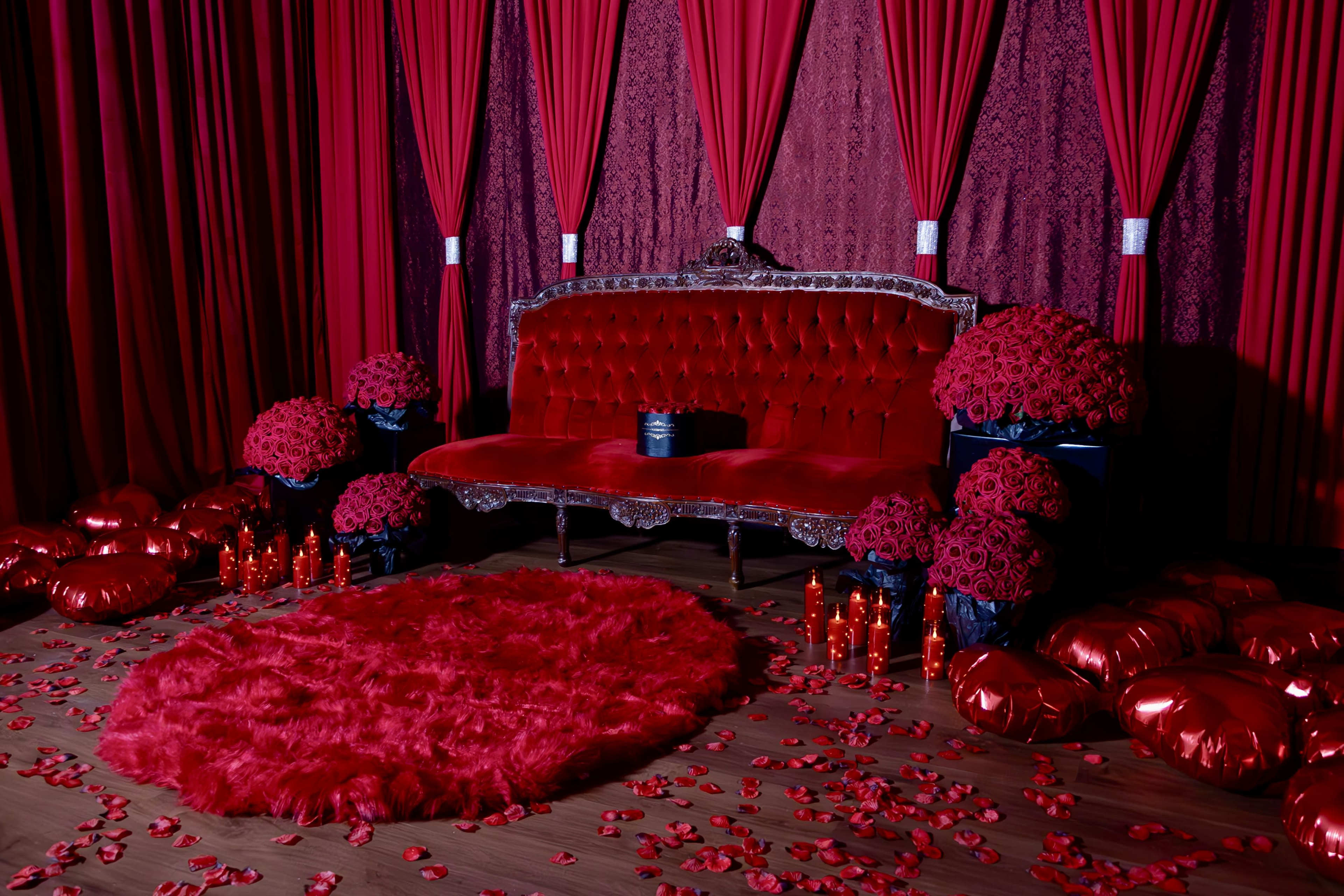 A lavish red room features a plush velvet sofa, surrounded by large bouquets of roses, candles, and scattered rose petals on the floor.