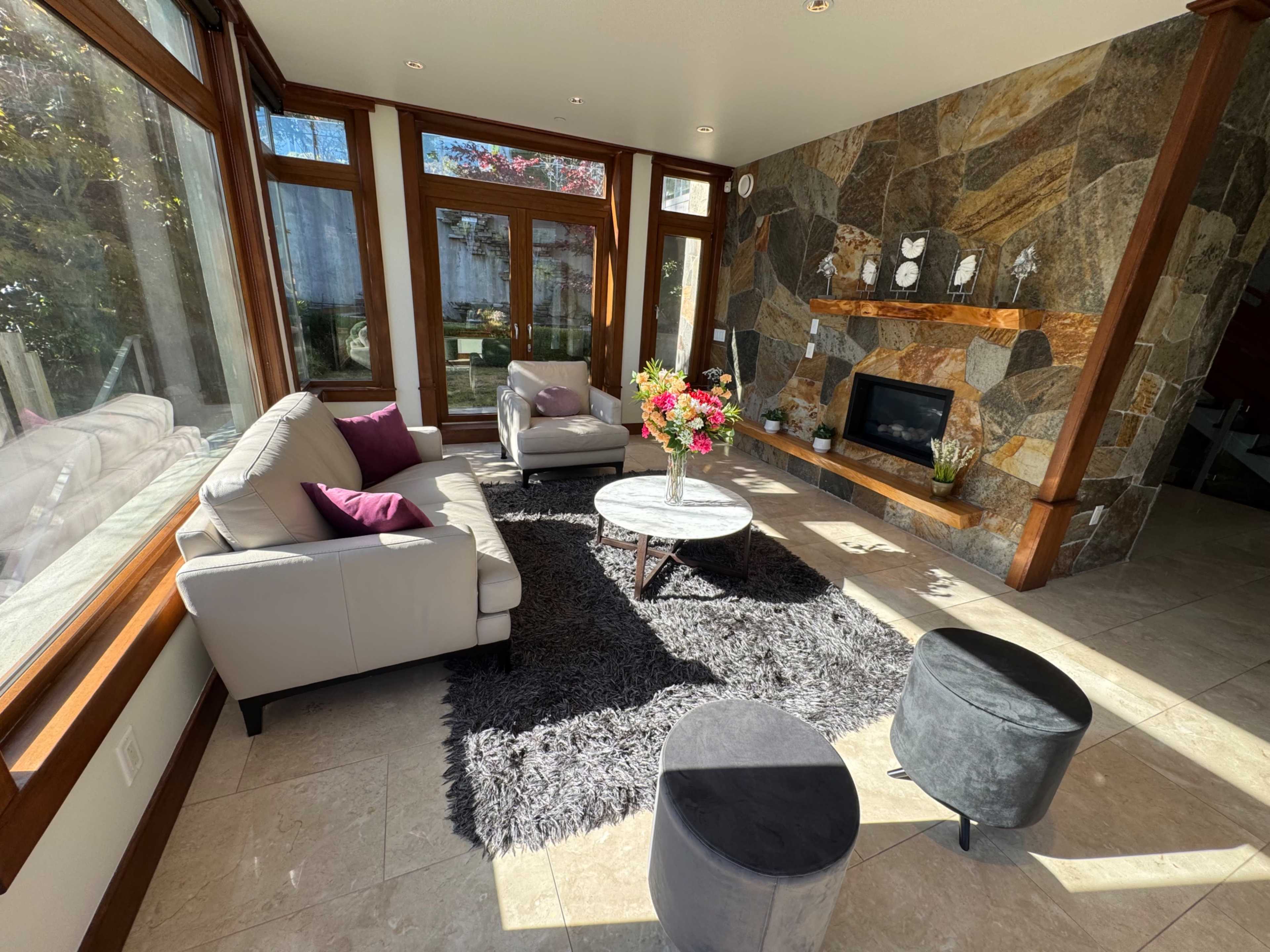 The image shows a well-lit living room with large windows, a stone feature wall, and modern furniture including a gray rug, a round coffee table, and two black stools.