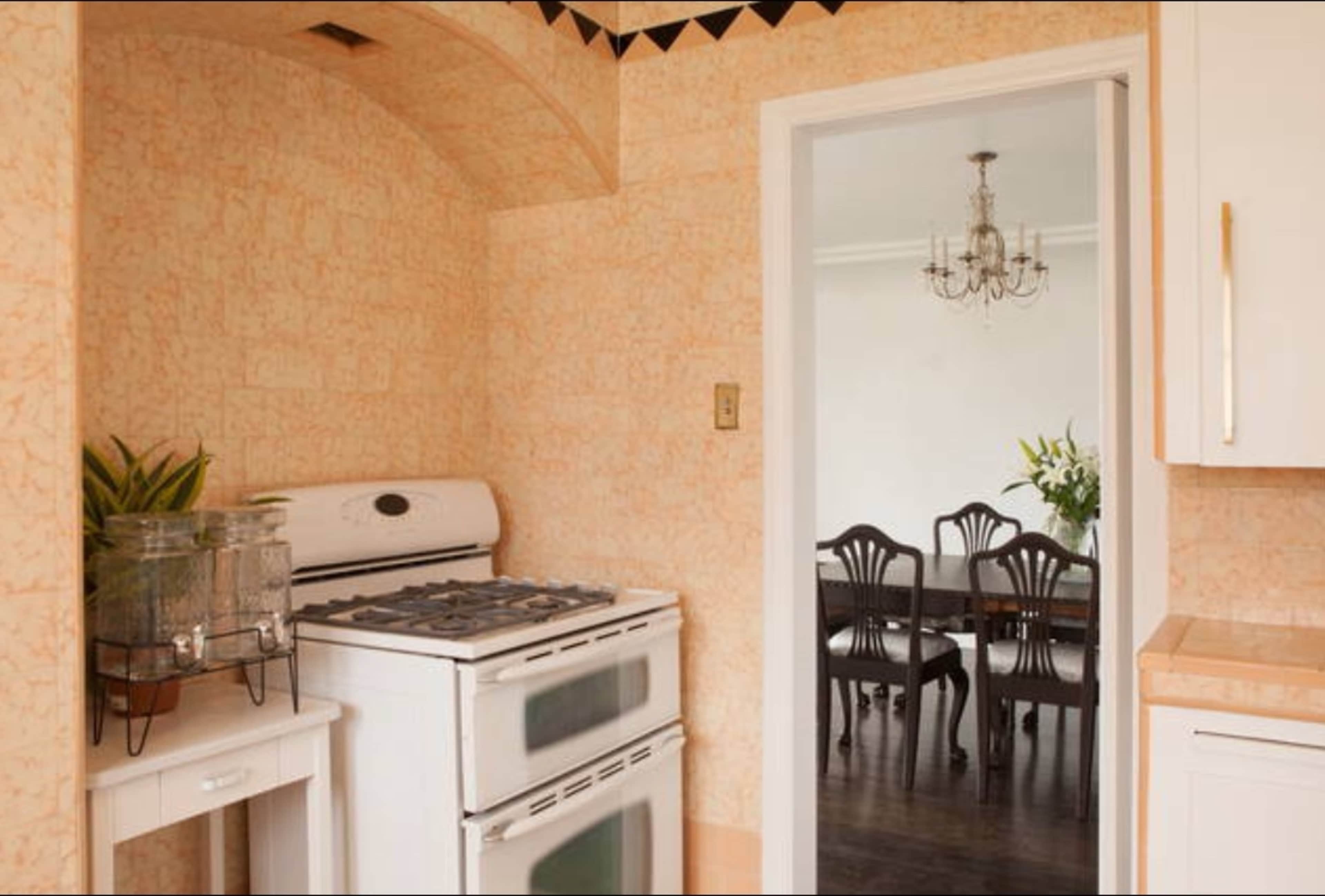 A kitchen features an upright white stove with a glass jug on a small table, leading into a dining area with a chandelier and wooden chairs.