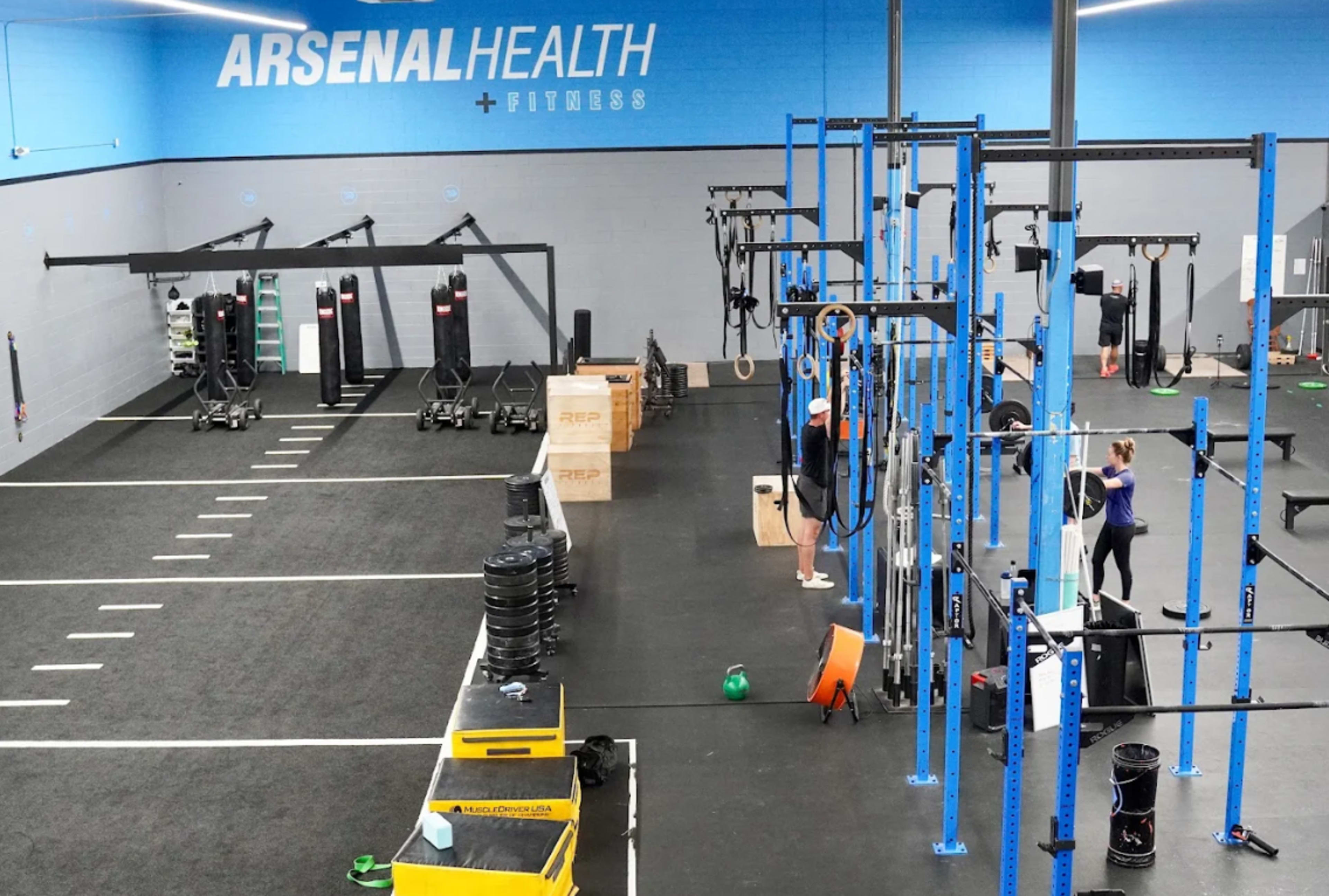 The image shows an indoor gym with various workout stations, equipment, and exercise mats, featuring blue and gray walls.