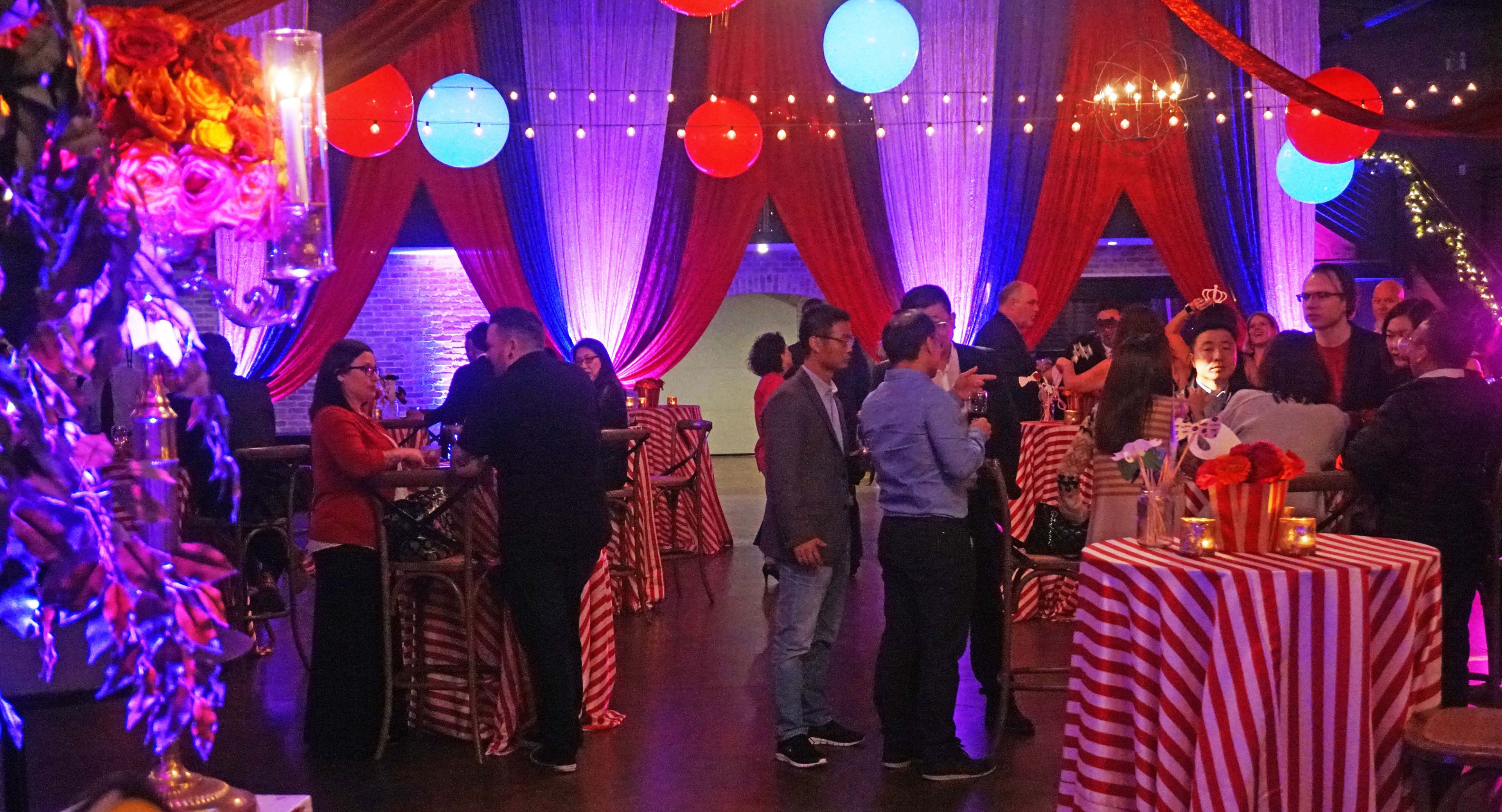 Guests socialize at tables with striped coverings in a festively decorated event space featuring colorful hanging lights and draped curtains.