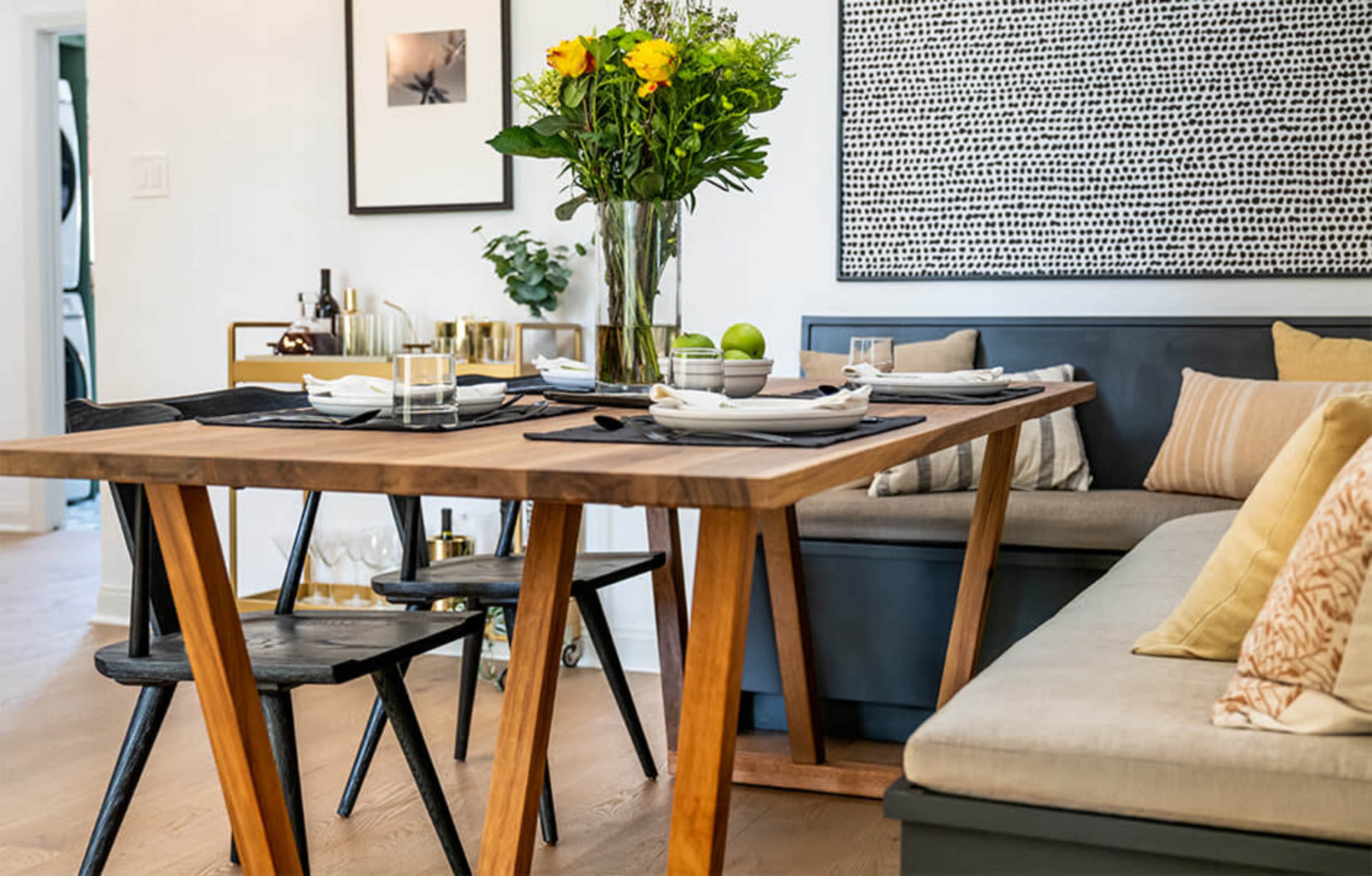A wooden dining table set with place settings, surrounded by chairs and a cushioned bench, displays a vase of flowers and decorative items in a modern dining area.