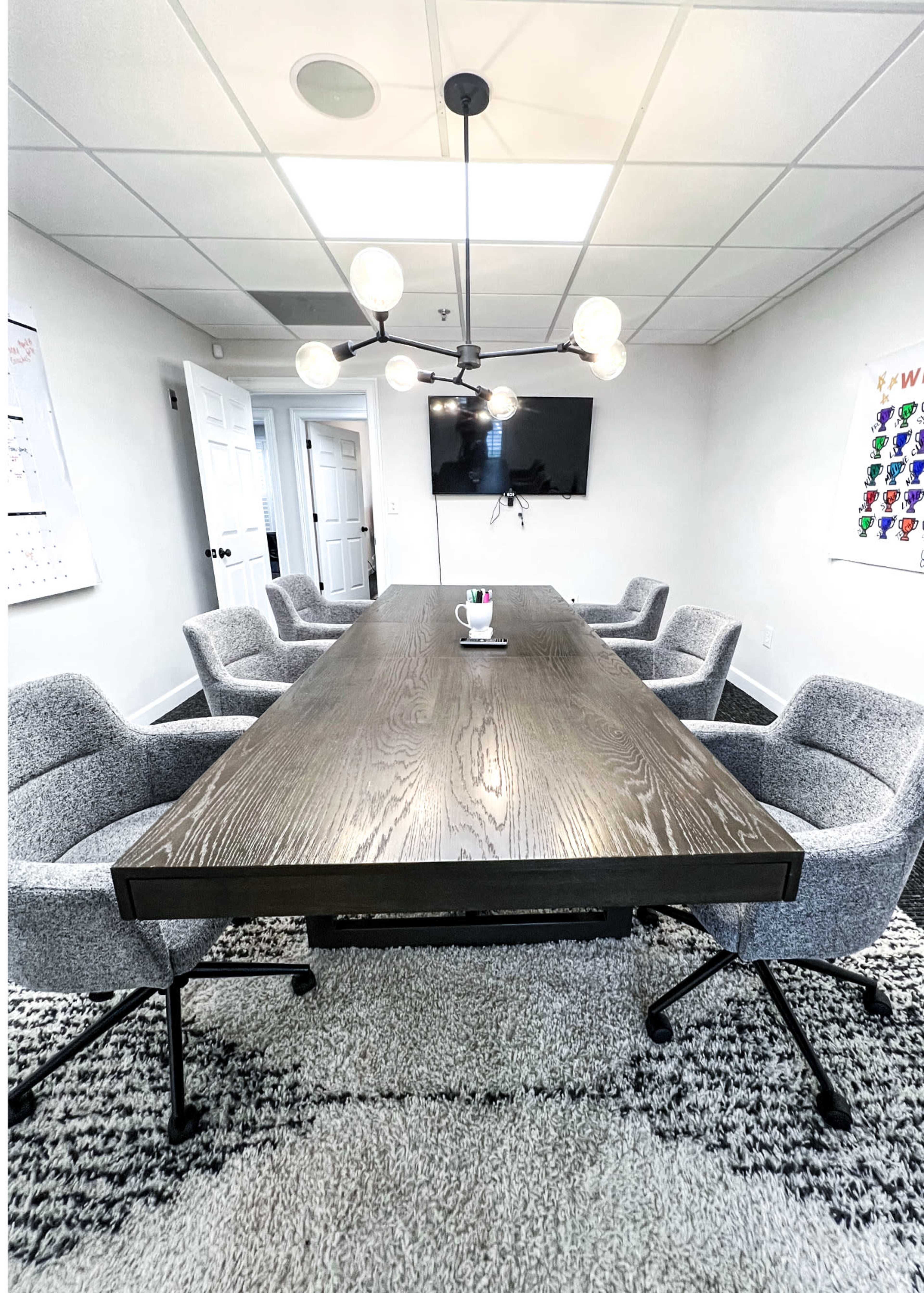 A modern conference room features a large wooden table surrounded by six upholstered chairs, with a wall-mounted screen and stylish light fixture overhead.