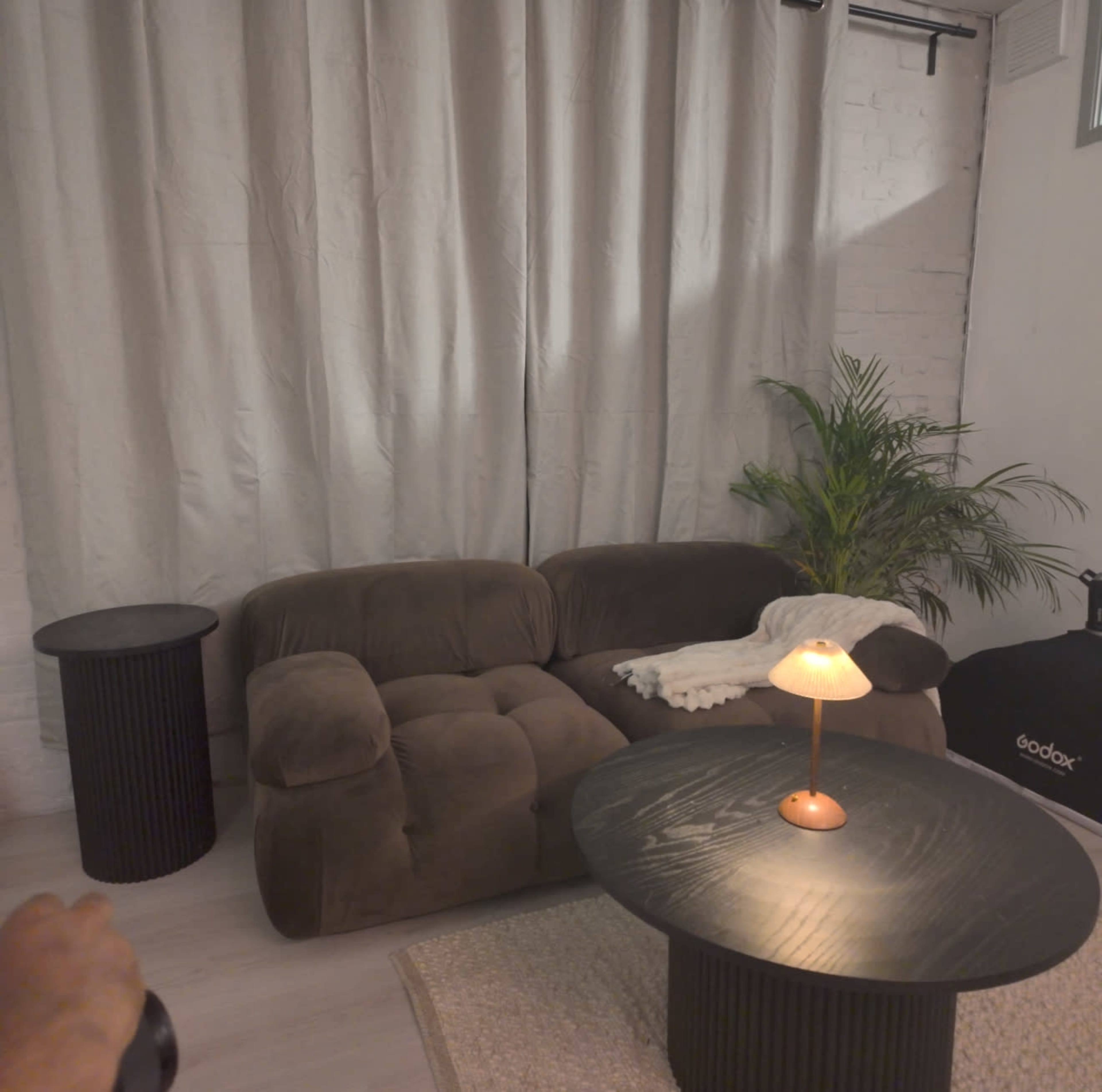 The image shows a cozy living room setup with a brown sectional couch, a small round table, a lamp, and a potted plant, all set against a backdrop of light curtains.