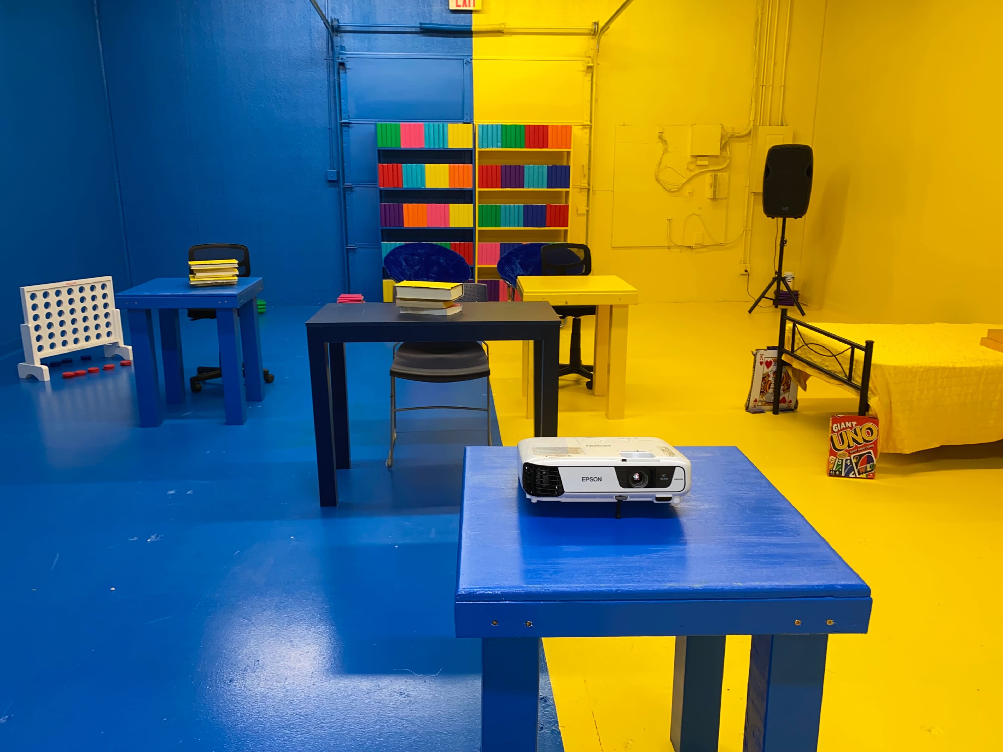 A colorful room divided by blue and yellow walls, featuring two tables with chairs, a projector on one table, and a shelf filled with colorful books.