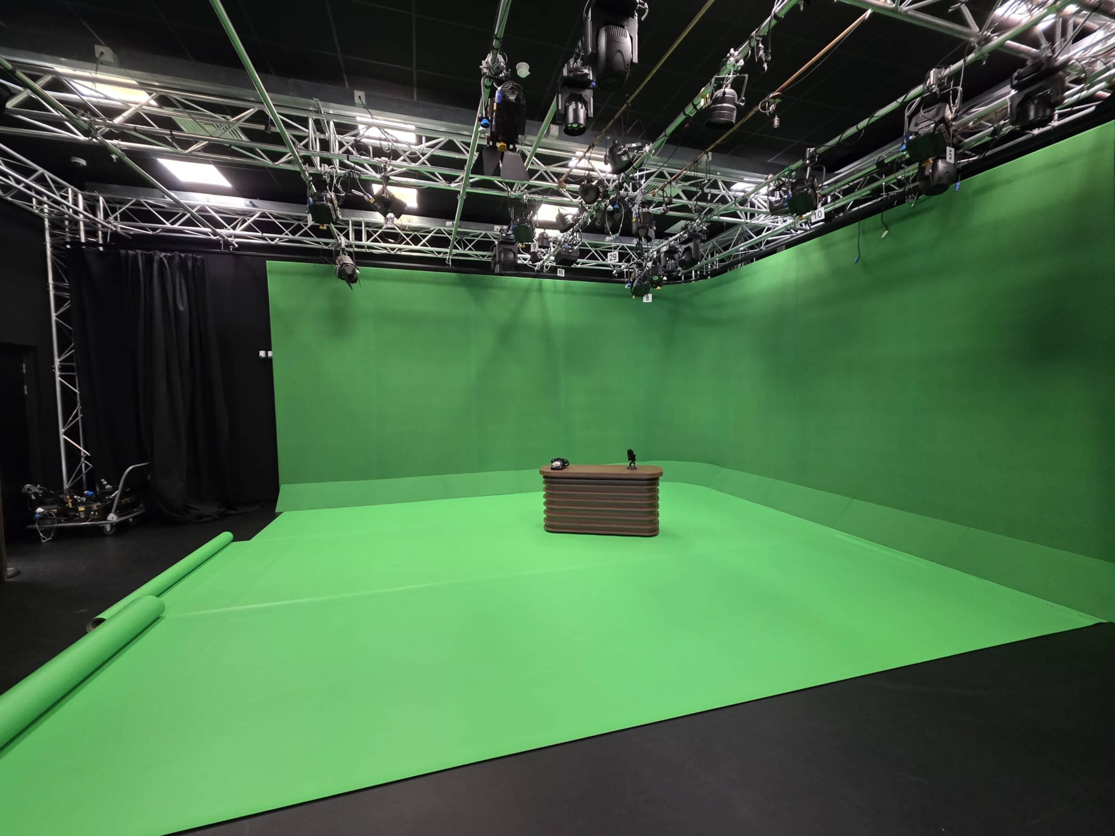The image shows a large green screen studio equipped with a wooden desk and various lighting fixtures.