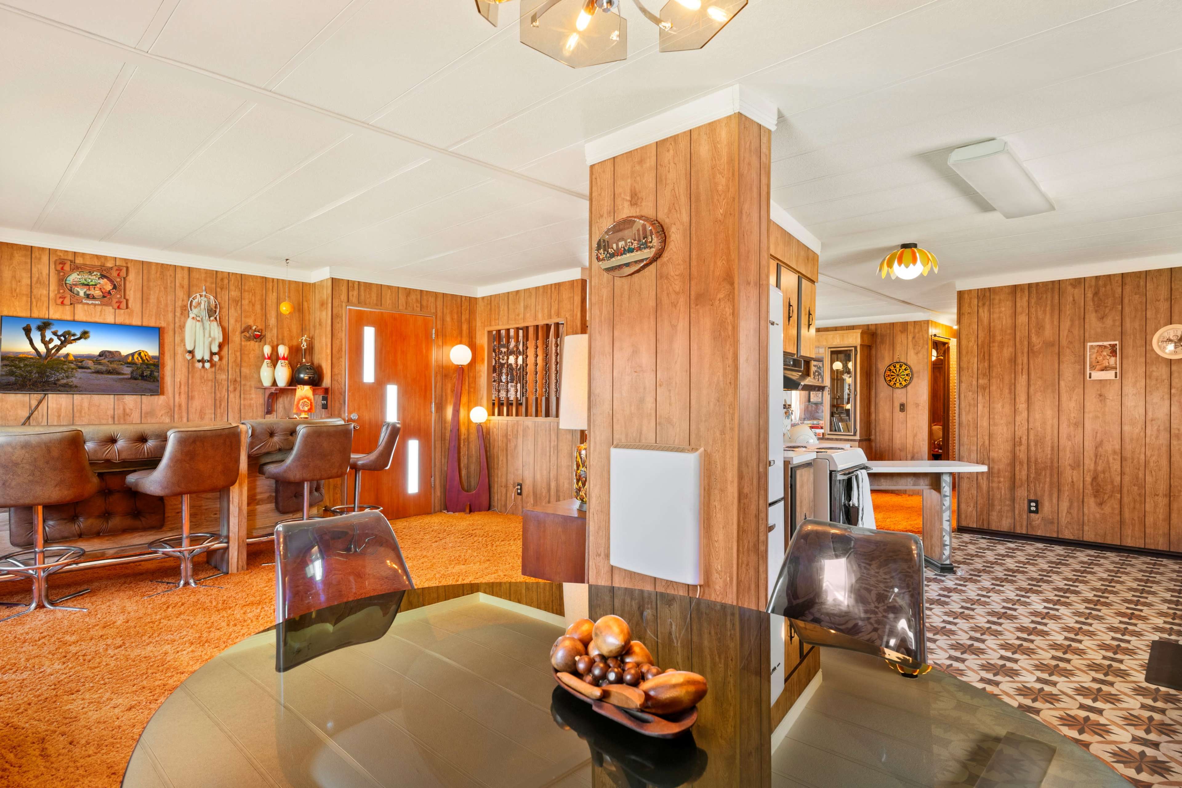 A retro-styled interior with wood-paneled walls, a circular glass dining table, and a bar area featuring brown stools and a textured floor.