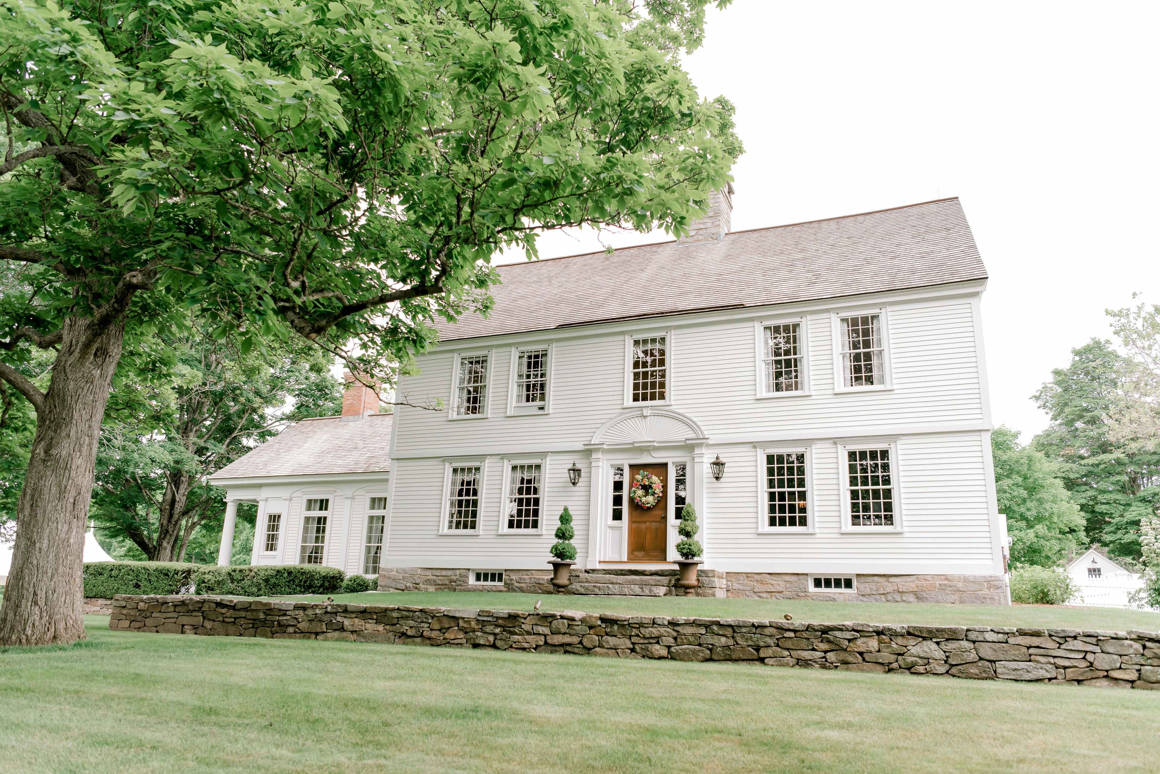 A two-story white colonial house sits on a grassy lawn surrounded by trees, with a stone wall in the foreground.