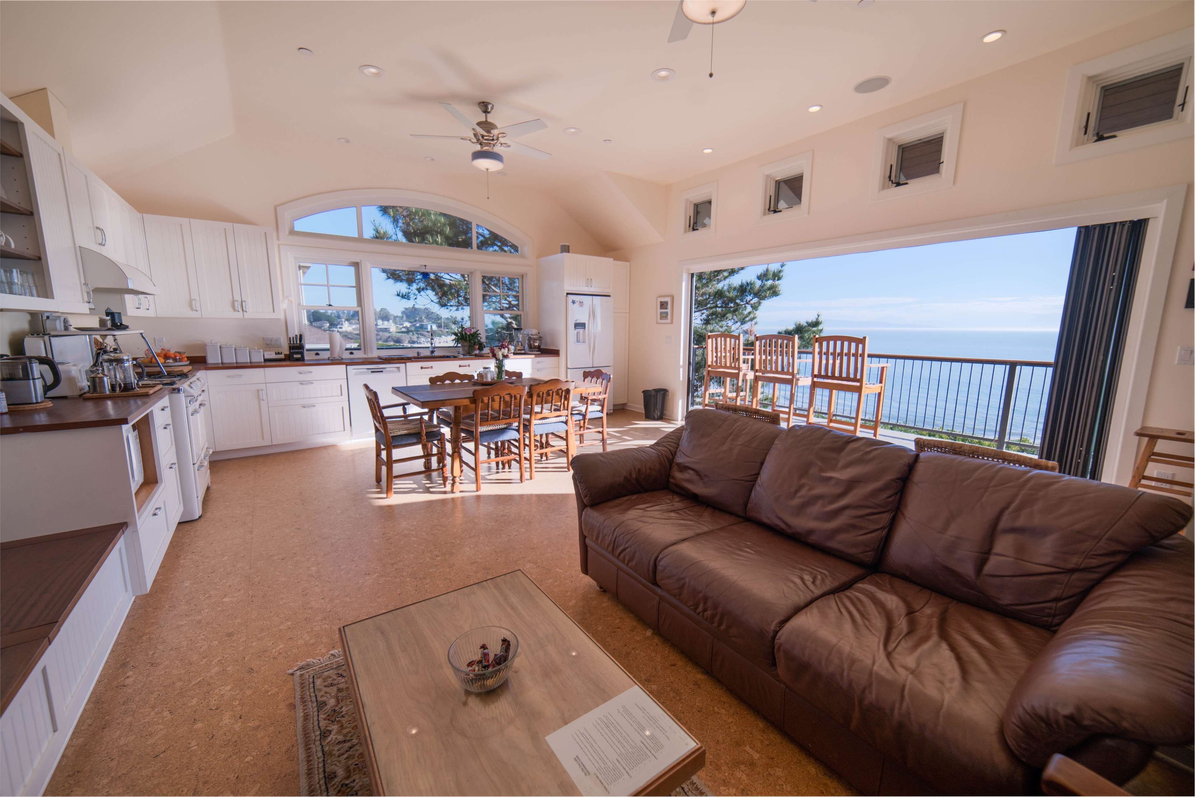 The image shows a kitchen and living area with a sofa, dining table, and large windows overlooking a water view.
