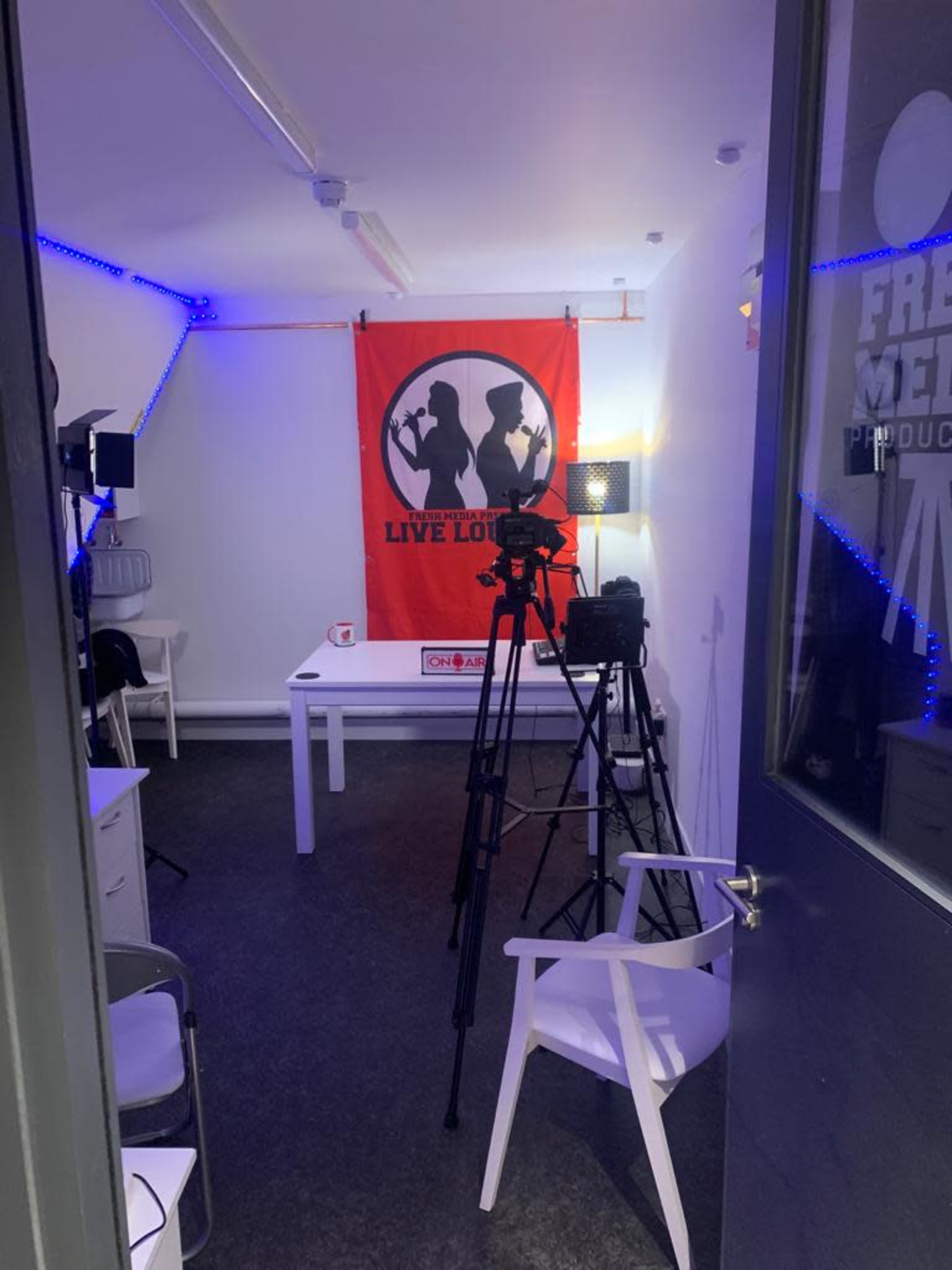 A video production room with a red backdrop, two tripods with cameras, and a table set up with lighting equipment.