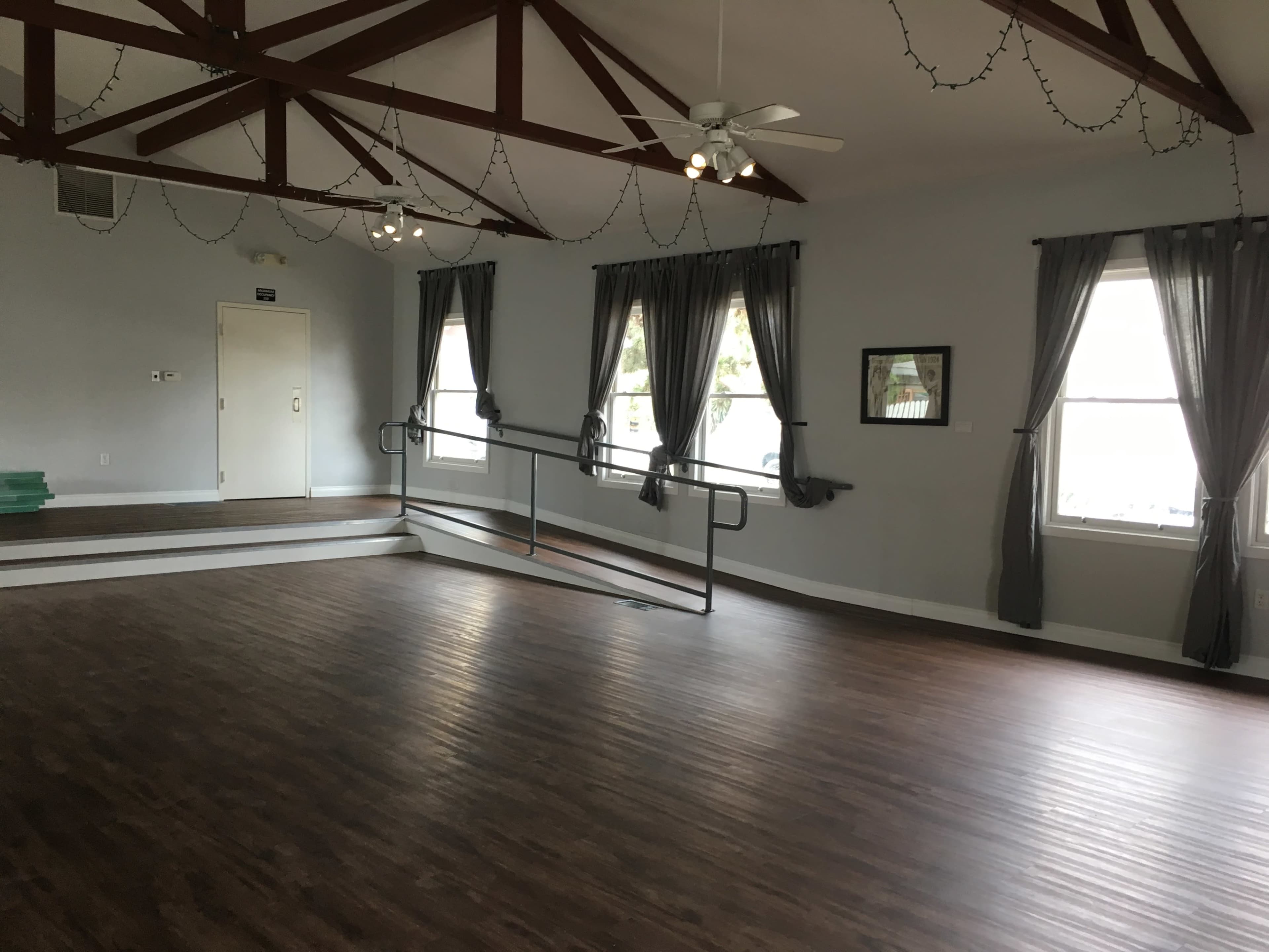 A spacious, empty room with wooden flooring, high ceilings supported by exposed beams, and large windows dressed with dark curtains.
