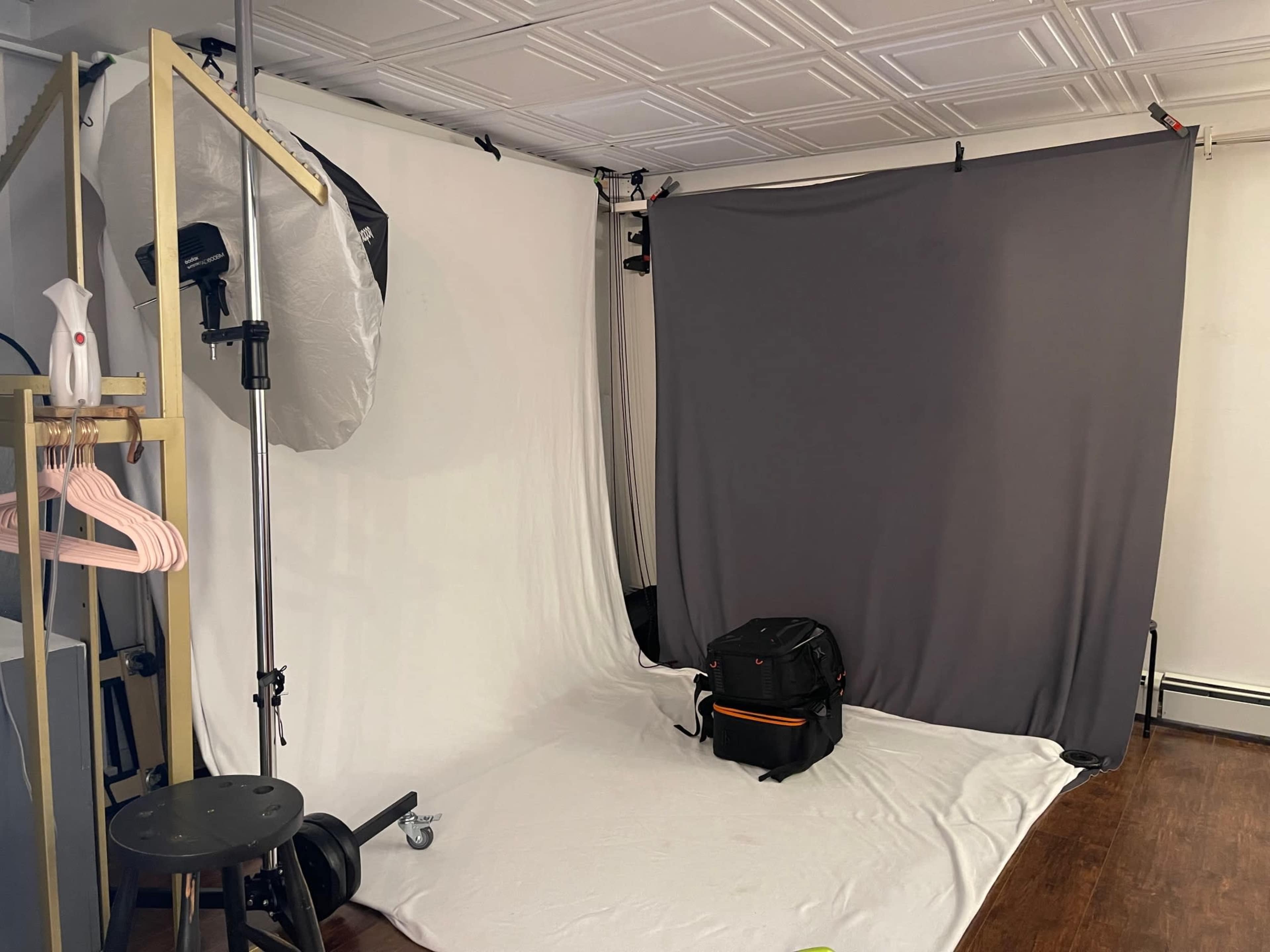 The image shows a photography studio setup with a gray backdrop, a white backdrop, a lighting umbrella, a stool, and a bag on the floor.