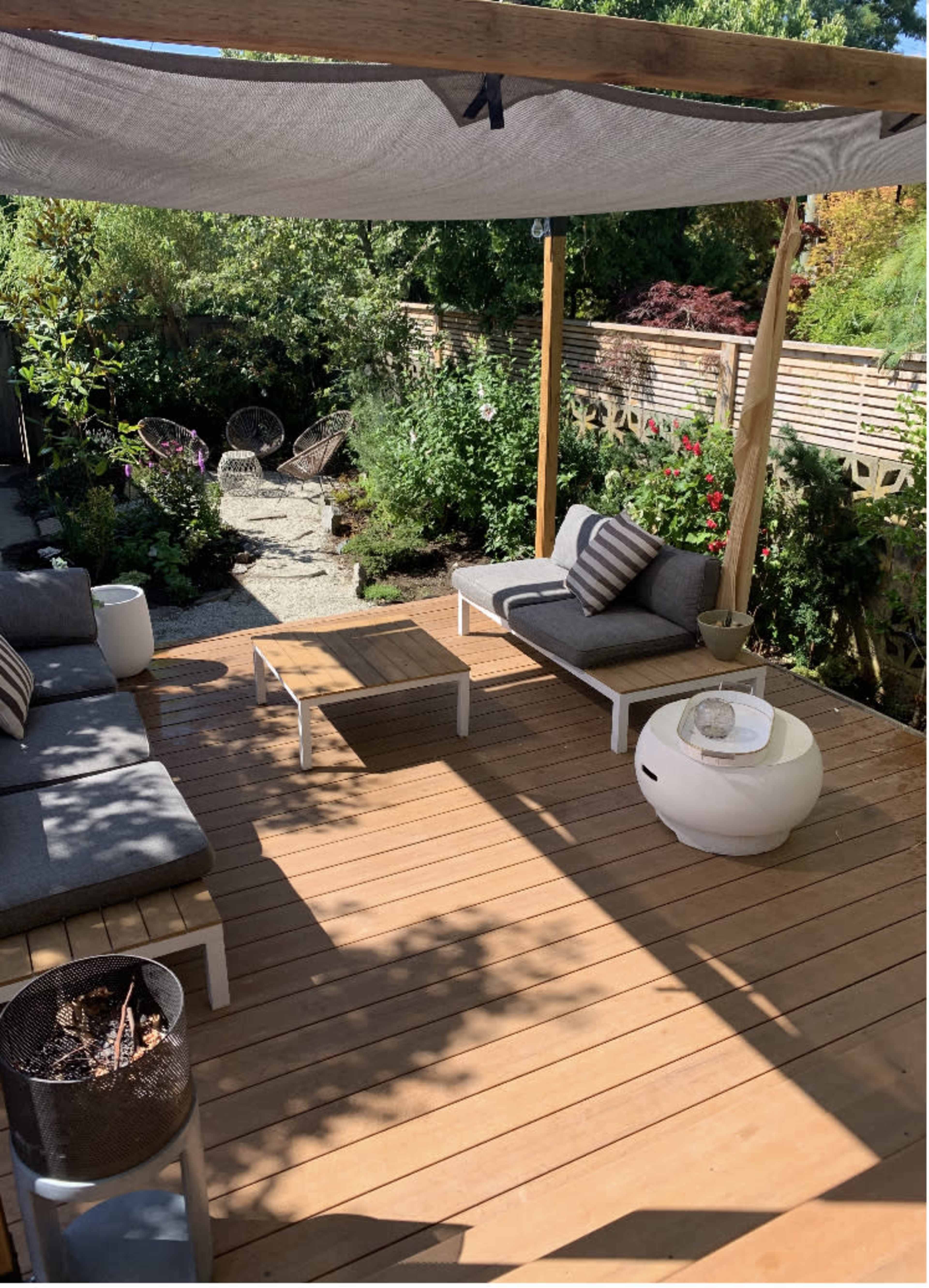 The image shows a wooden patio area with two lounge chairs, a coffee table, and a shaded seating arrangement surrounded by greenery and stone pathways.