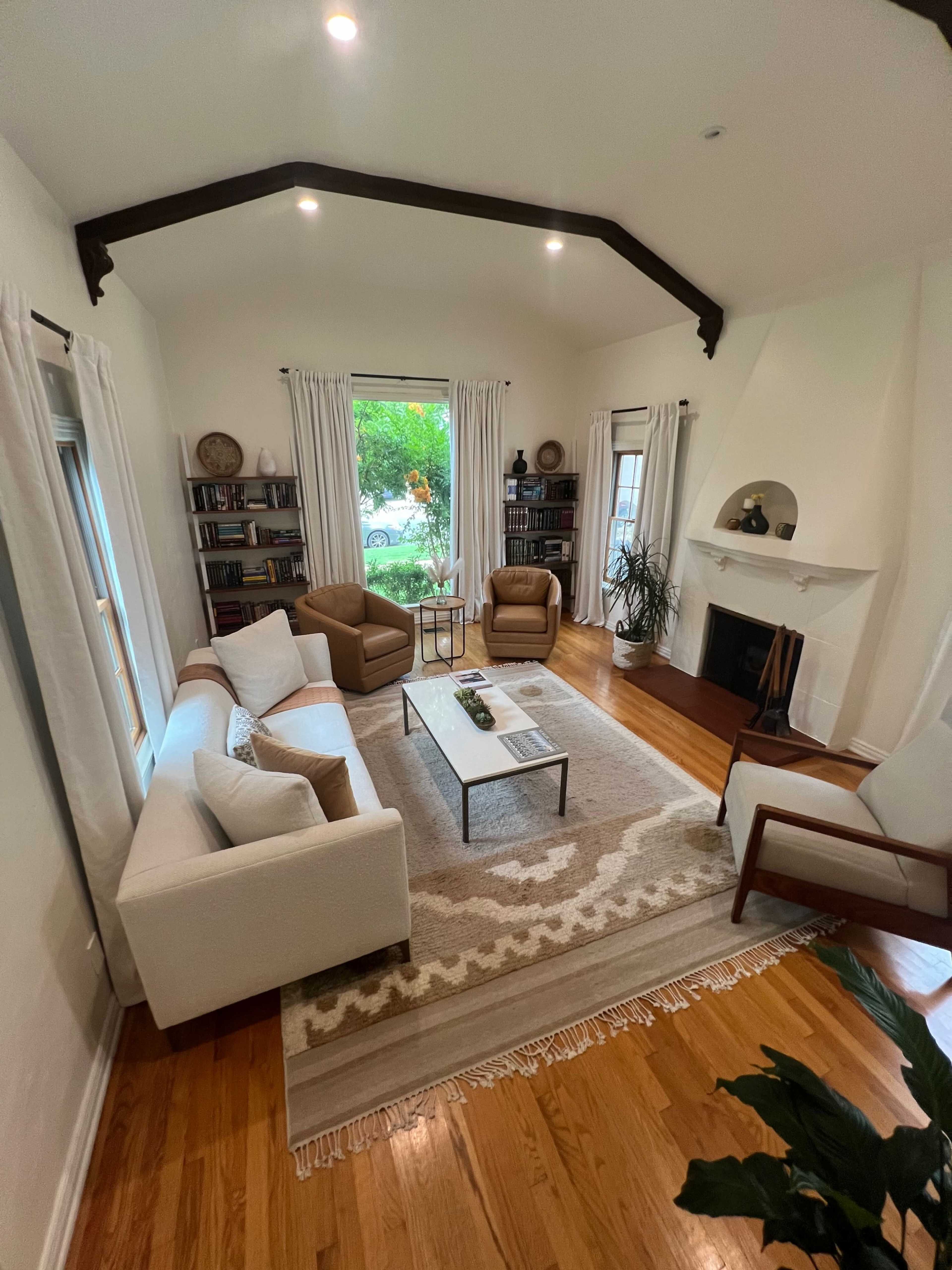 A well-furnished living room with two armchairs, a sofa, a coffee table, a fireplace, and bookshelves along the walls, all illuminated by natural light.
