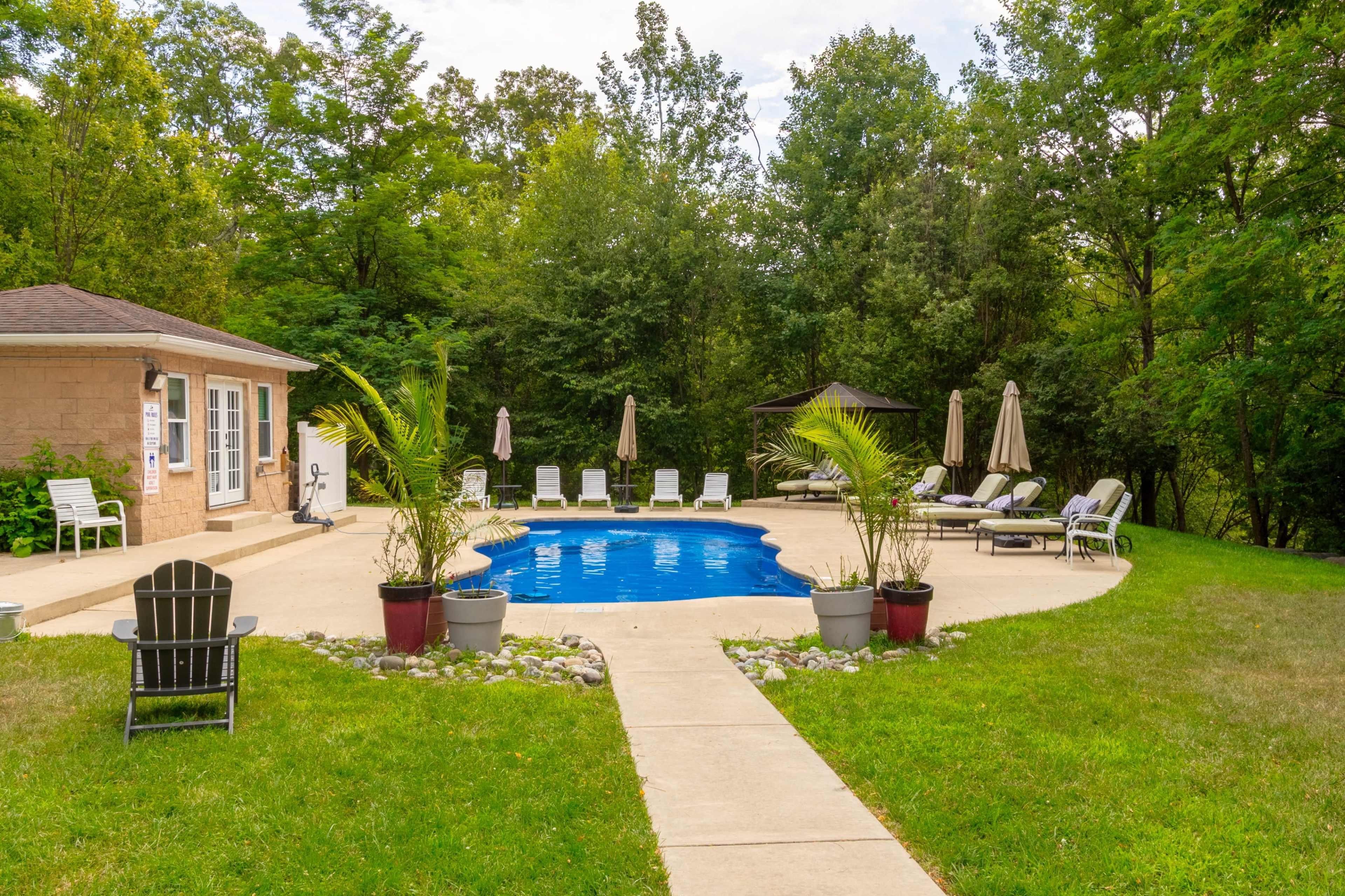 The image shows a swimming pool surrounded by lounge chairs and umbrellas, set in a landscaped area with trees in the background.