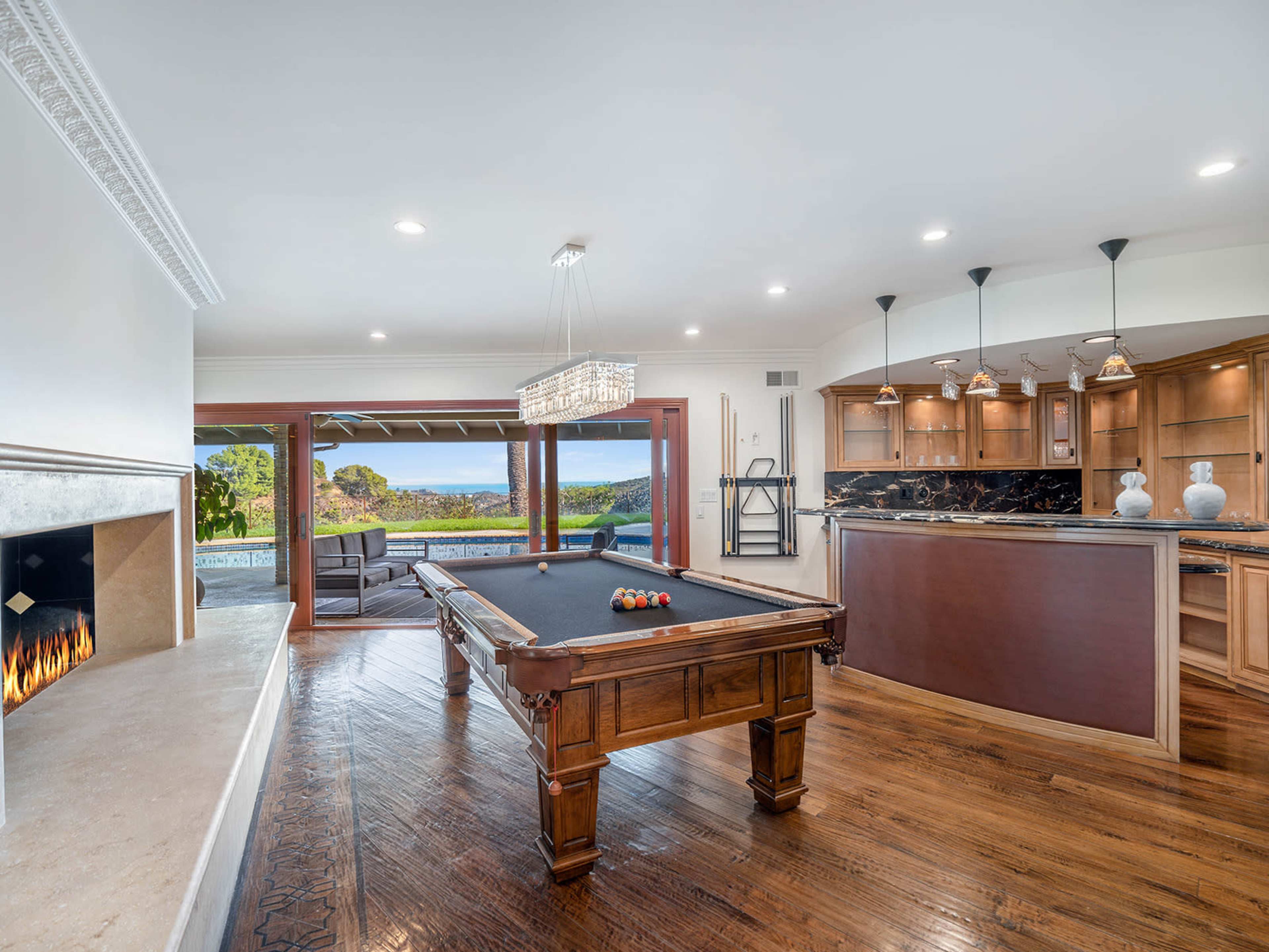 The image shows a spacious interior with a billiard table, a modern kitchen area, and large windows offering a view of the outdoors.