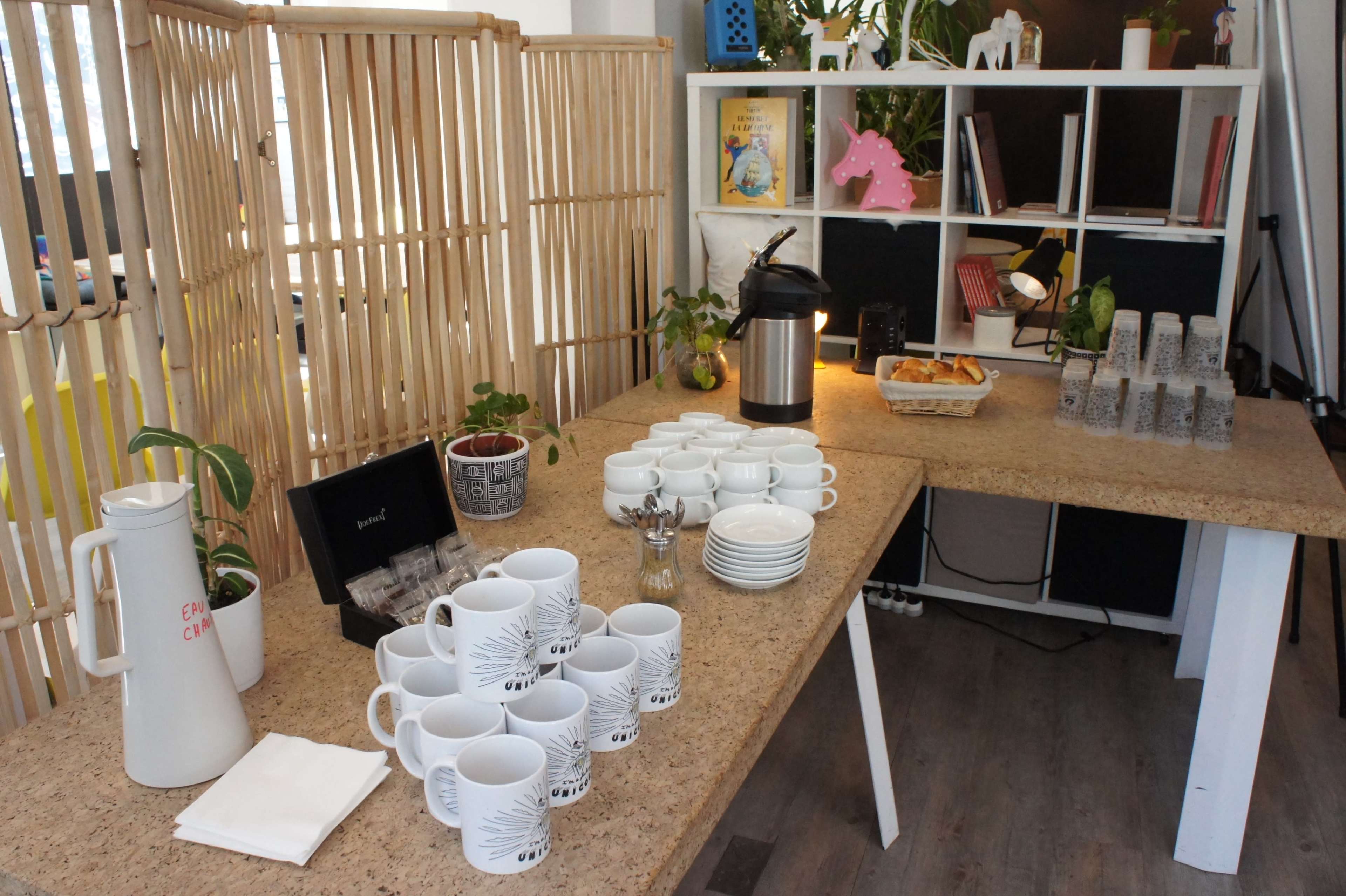A table is set up with various white mugs, plates, a coffee pot, and snacks, surrounded by plants and books on shelves.