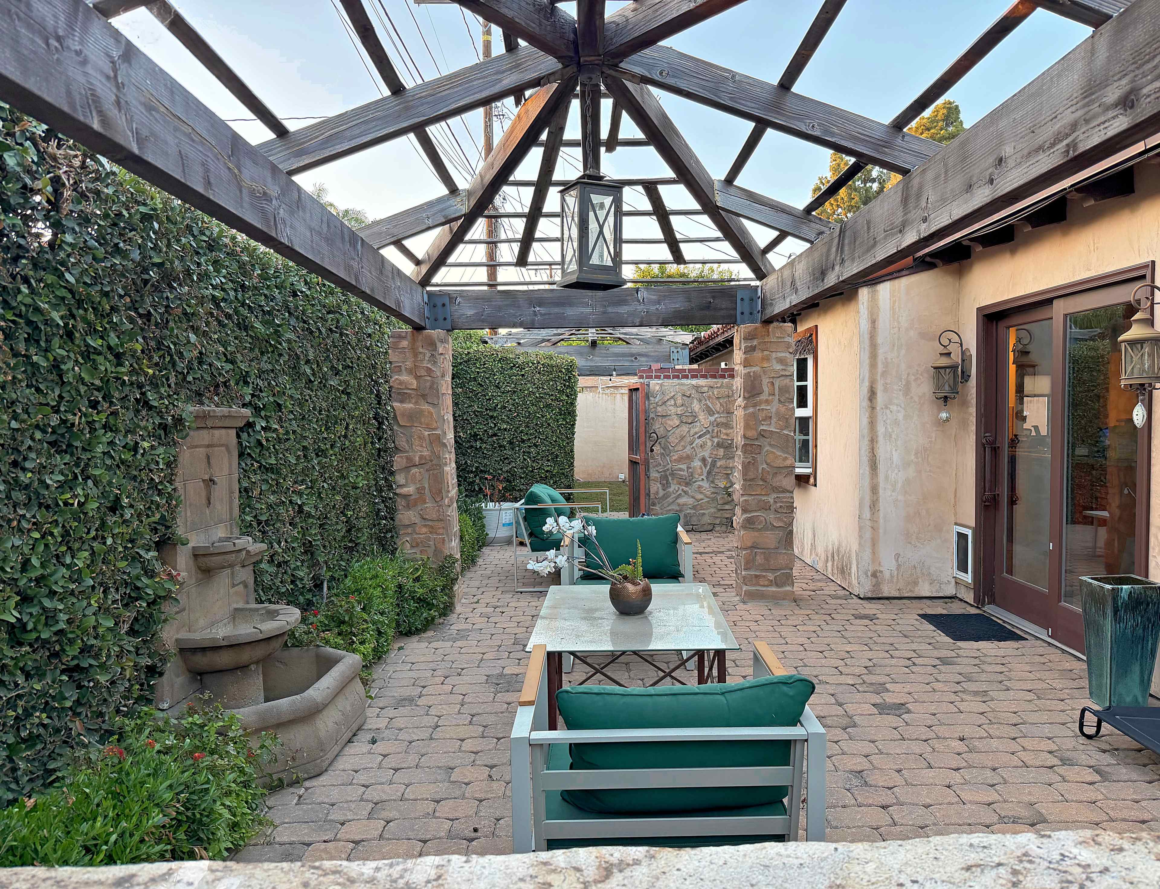The image shows a patio area with a stone wall, a table surrounded by chairs, and a decorative fountain under a wooden pergola.