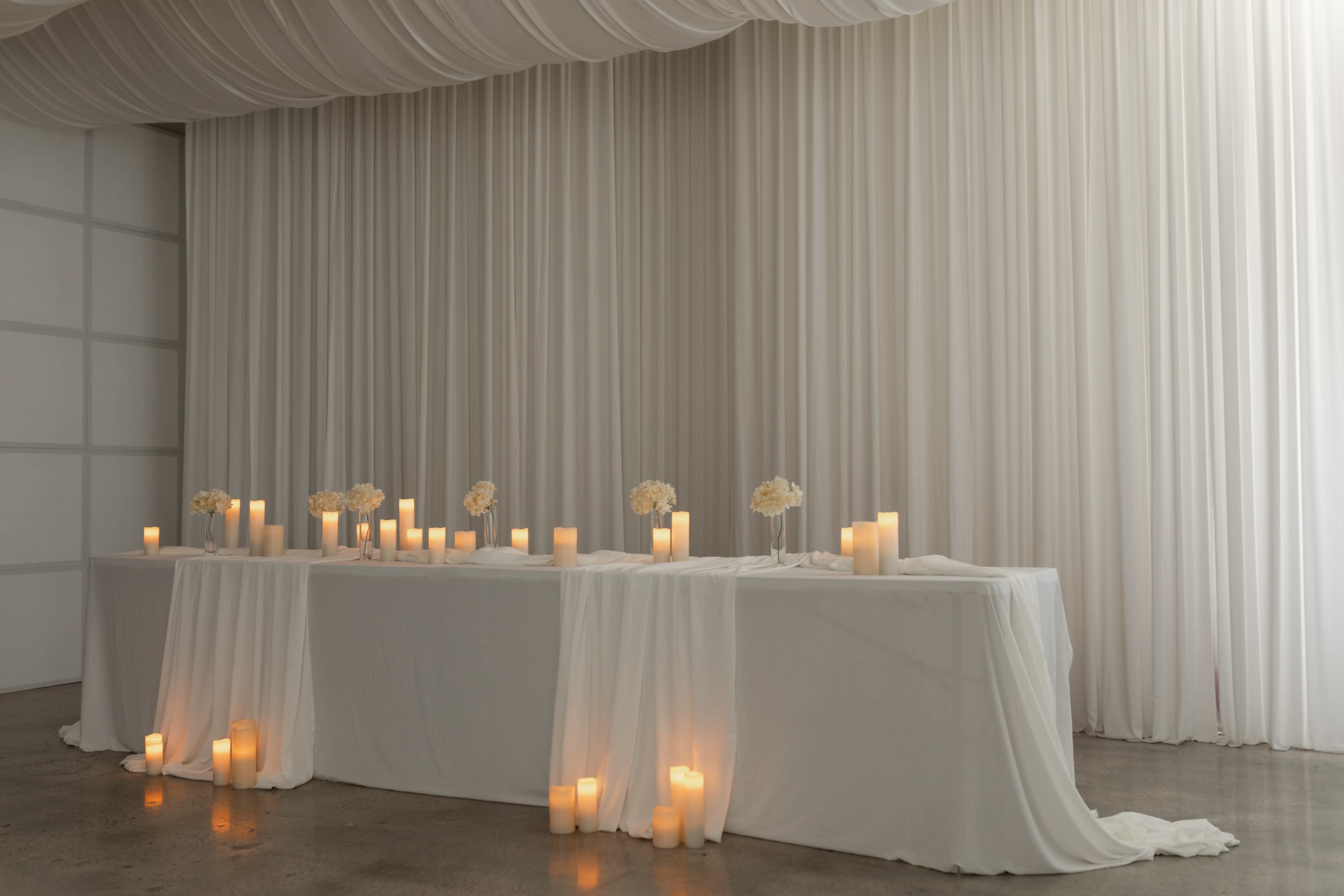 A long table is elegantly set with white drapes and adorned with candles and flower arrangements in a softly lit room.