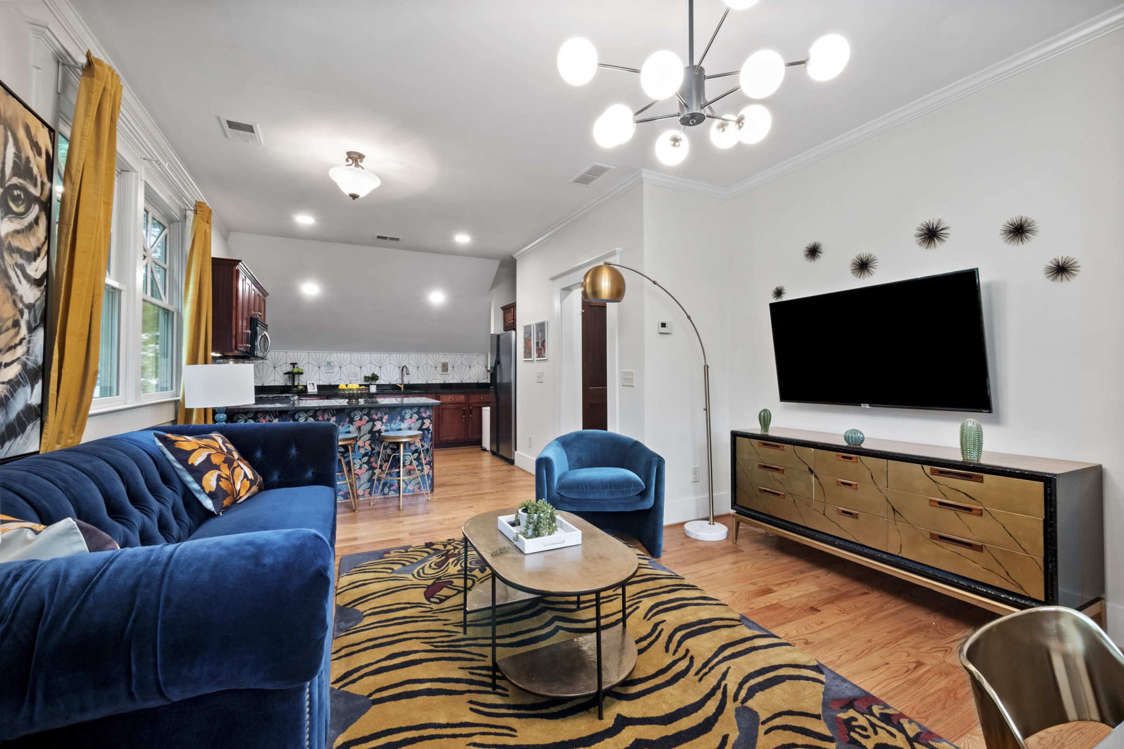 A modern living room featuring a blue velvet sofa, a gold-accented console table, and a large flat-screen TV mounted on the wall, complemented by a decorative yellow and black rug.