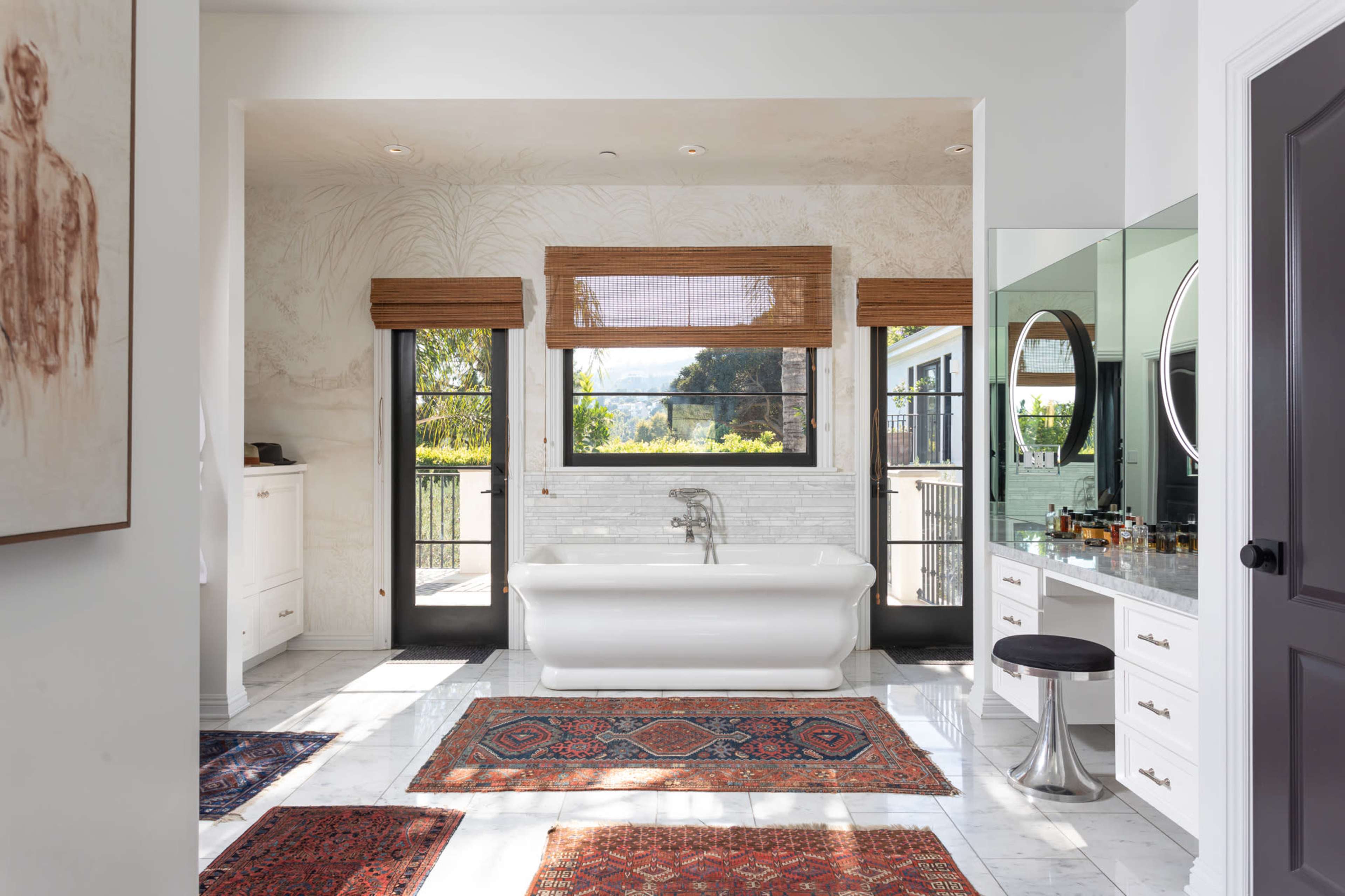 A spacious bathroom features a freestanding white bathtub, large windows with bamboo shades, and marble flooring accented by patterned rugs.