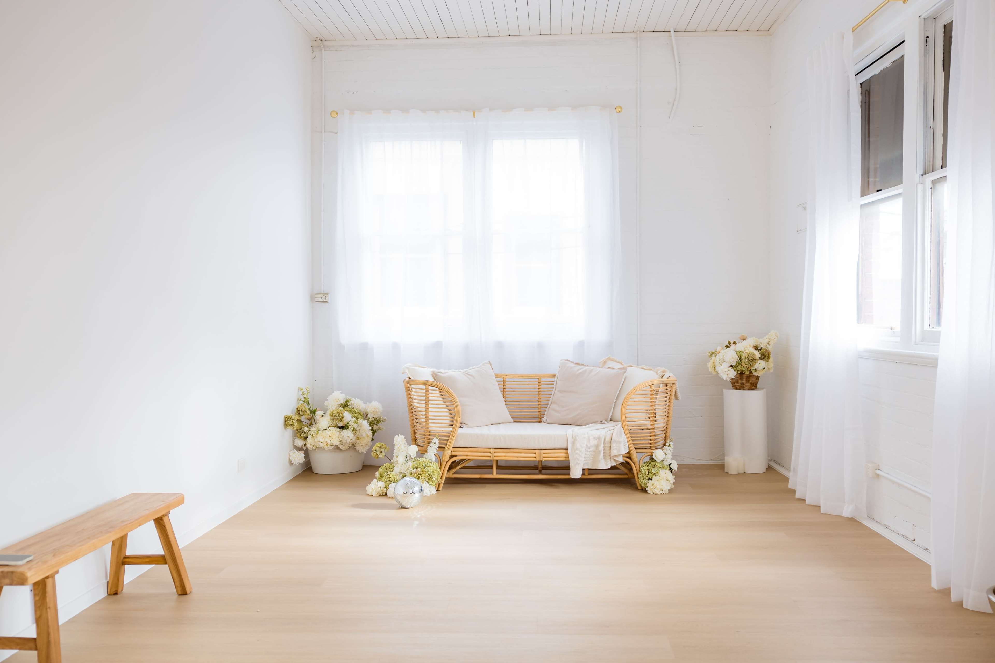 The image shows a bright, minimalistic room with a rattan sofa adorned with cushions and white flowers, and large windows allowing natural light to enter.