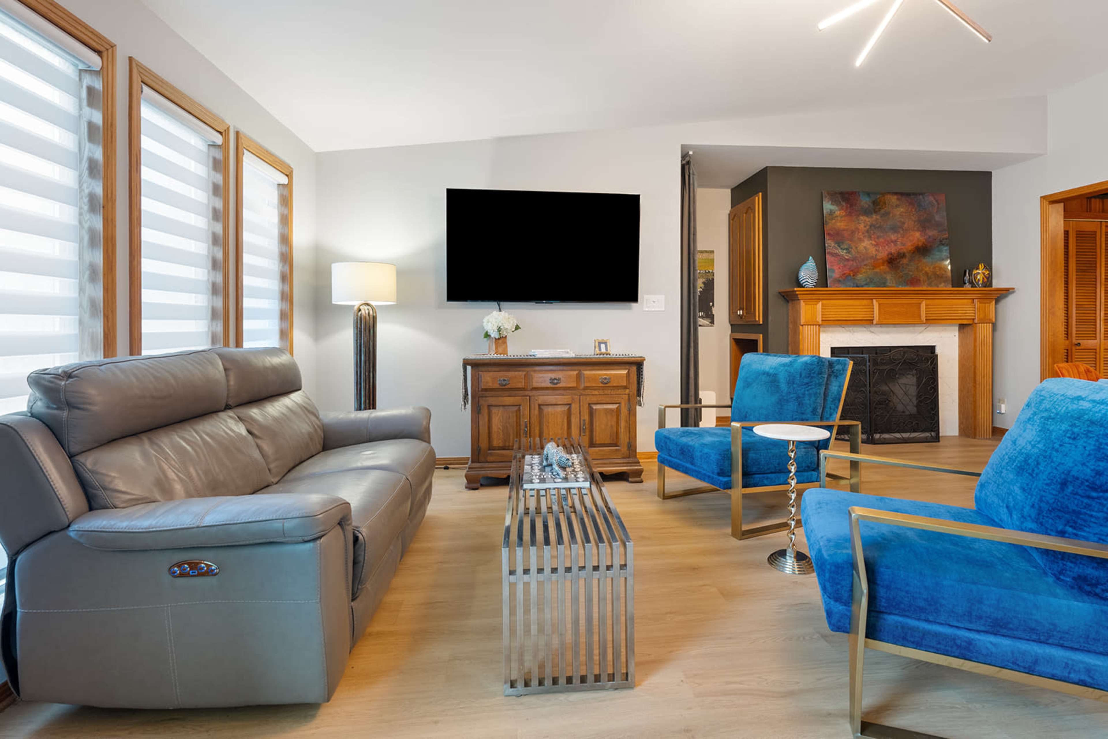 The image shows a modern living room with a leather sofa, two blue accent chairs, a wooden cabinet, a fireplace, and a mounted television.